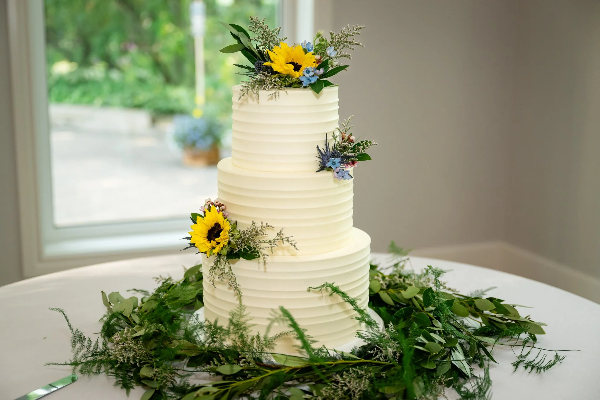 Three-tiered white wedding cake decorated with yellow sunflowers, blue flowers, and greenery, placed on a table with green foliage around the base.