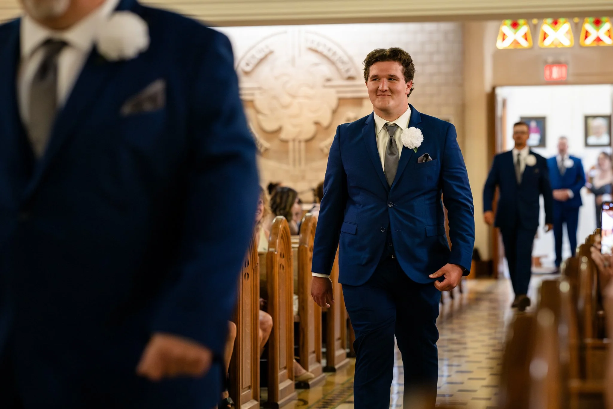 A man in a blue suit walking down the aisle of a church during a wedding ceremony, with other men in suits in the background.