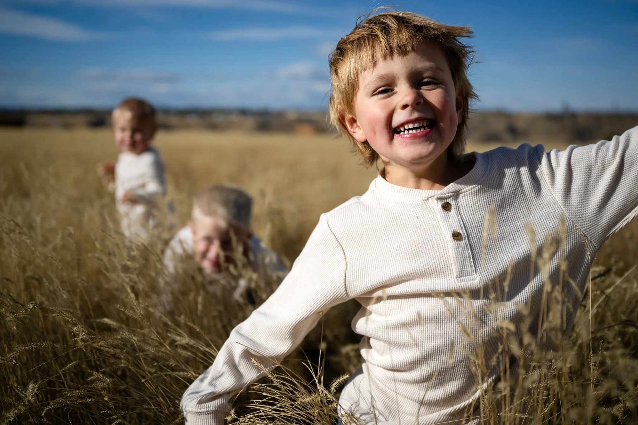 Smiling young boy with red hair in a white shirt standing in a field of tall, golden grass with three other children in the background.