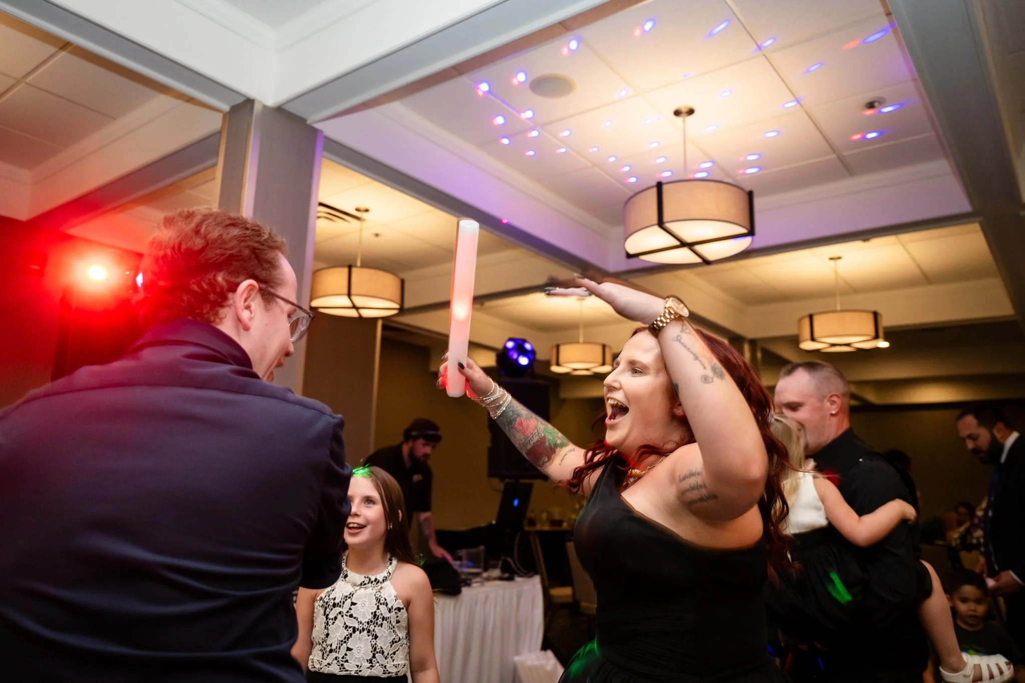 People dancing and celebrating at a party in a decorated indoor venue, with colorful lights and a DJ in the background.