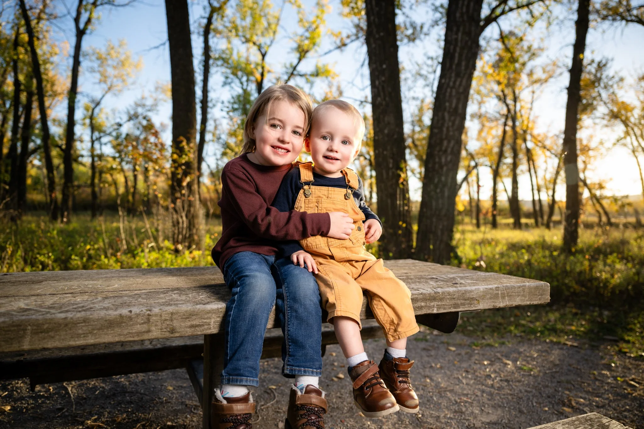Two children sitting on a wooden bench outdoors in a forest during autumn, smiling at the camera.