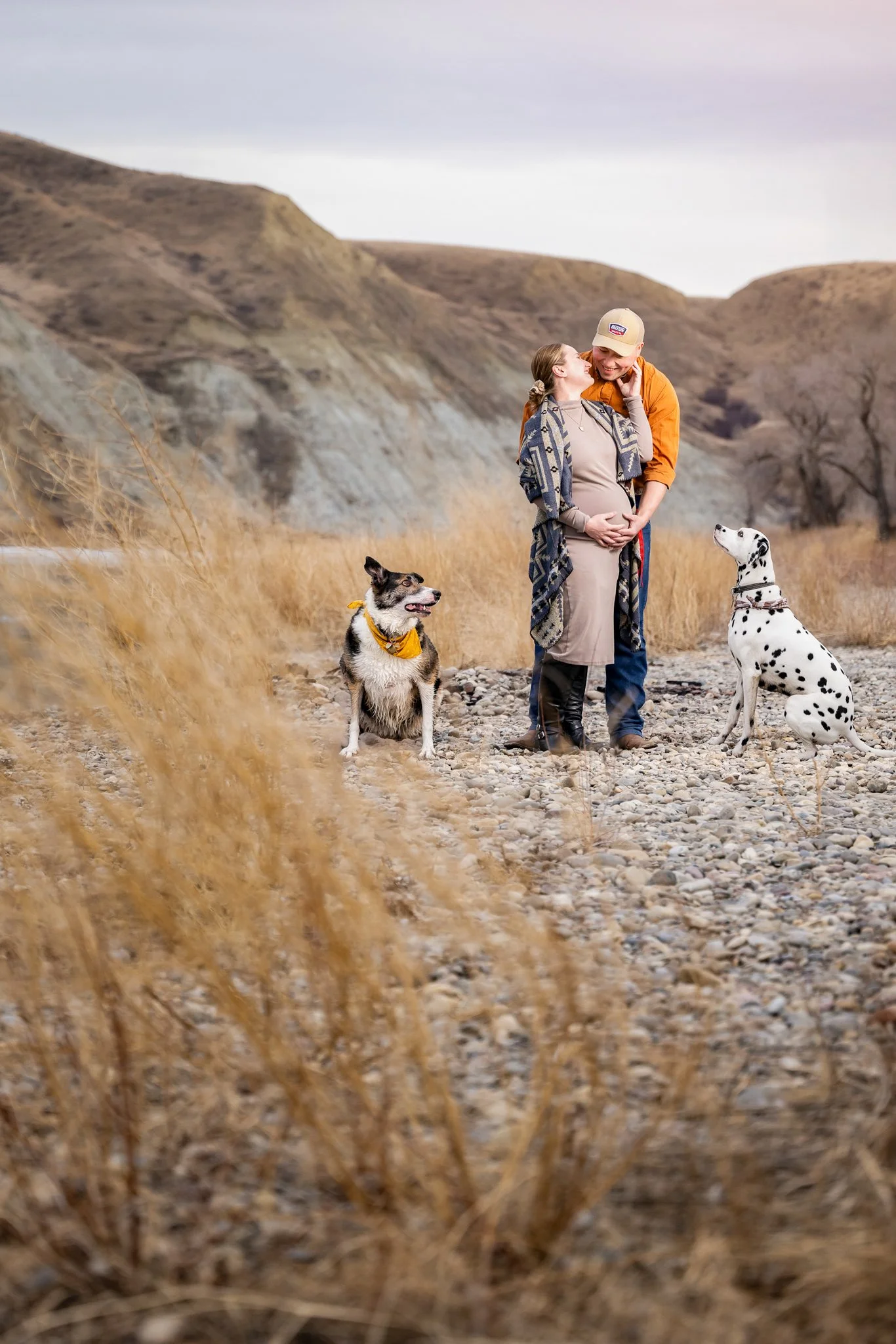 A pregnant woman and a man sharing a kiss outdoors with two dogs, one sitting and one standing, in a rocky, grassy landscape with hills in the background.