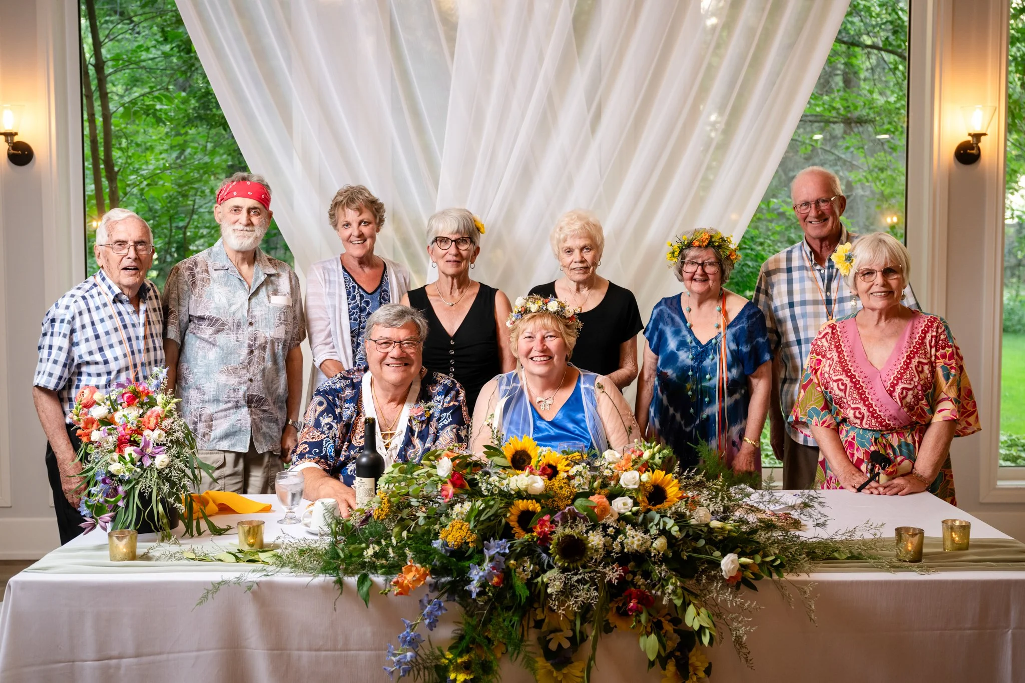 A group of elderly people celebrating at a wedding reception, with two women wearing flower crowns, seated at a decorated table with flowers and bottles, in front of a large window with white drapes and greenery outside.