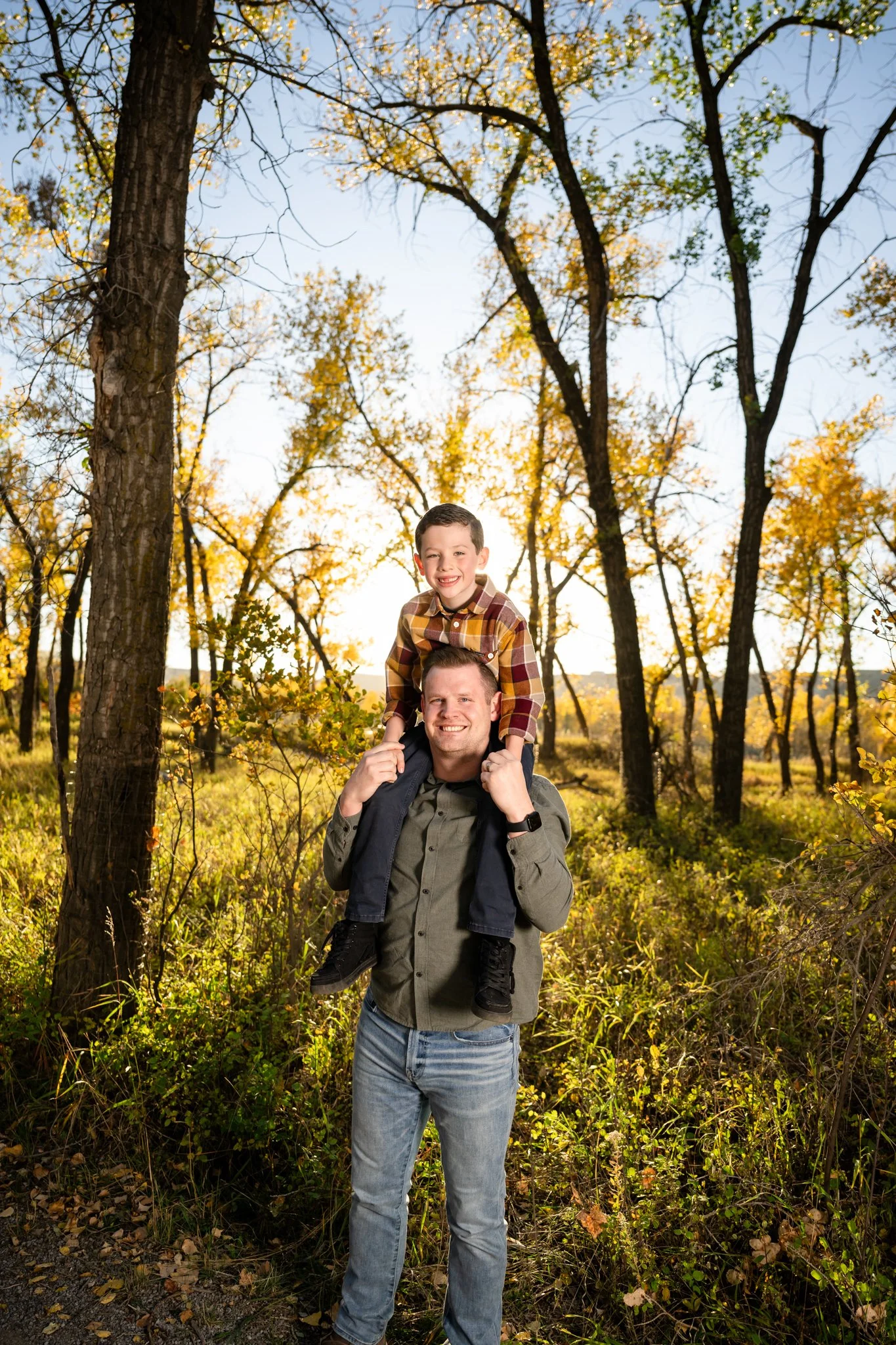 A man carrying a young boy on his shoulders, smiling, in a wooded area during fall with yellow leaves.