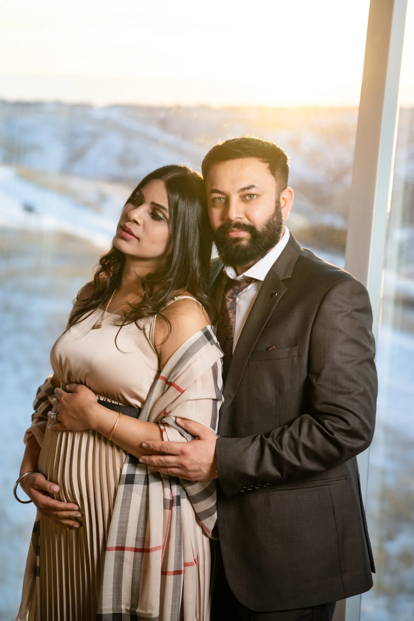 A pregnant woman standing next to a man, both dressed formally, with the woman wearing a beige dress and the man in a dark suit. They are inside near a large window with a winter landscape outside at sunset.