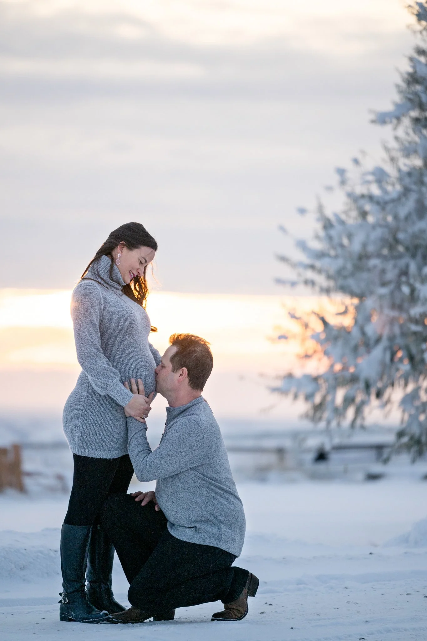 A man kneeling and kissing a pregnant woman on her belly outdoors in a snowy landscape during sunset.