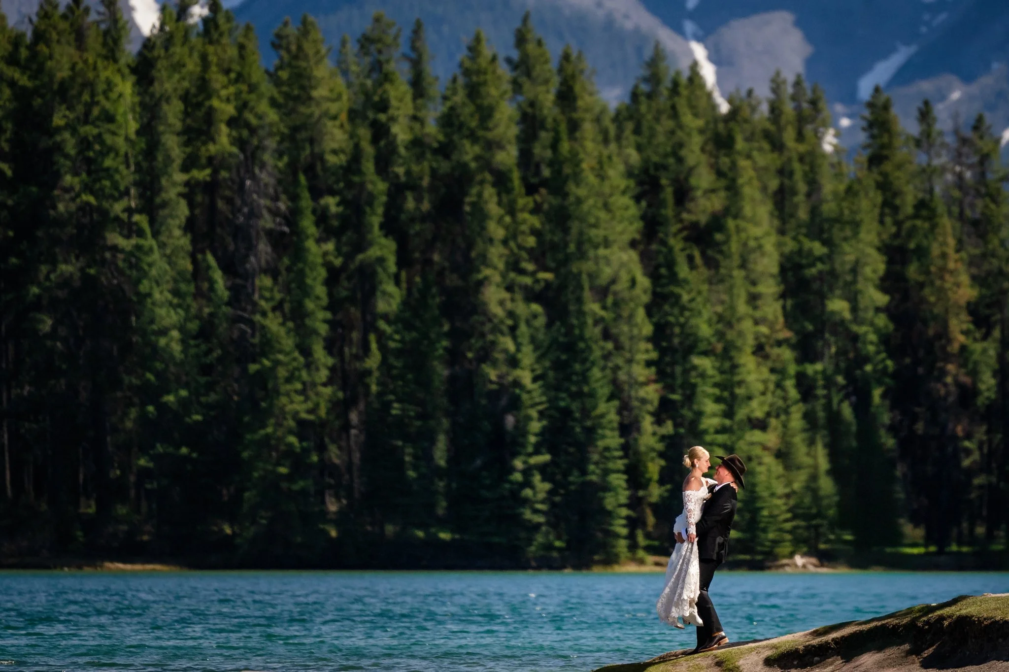 A bride and groom standing on a rock by a lake, with a forest of tall pine trees and mountains in the background, during a romantic outdoor wedding.