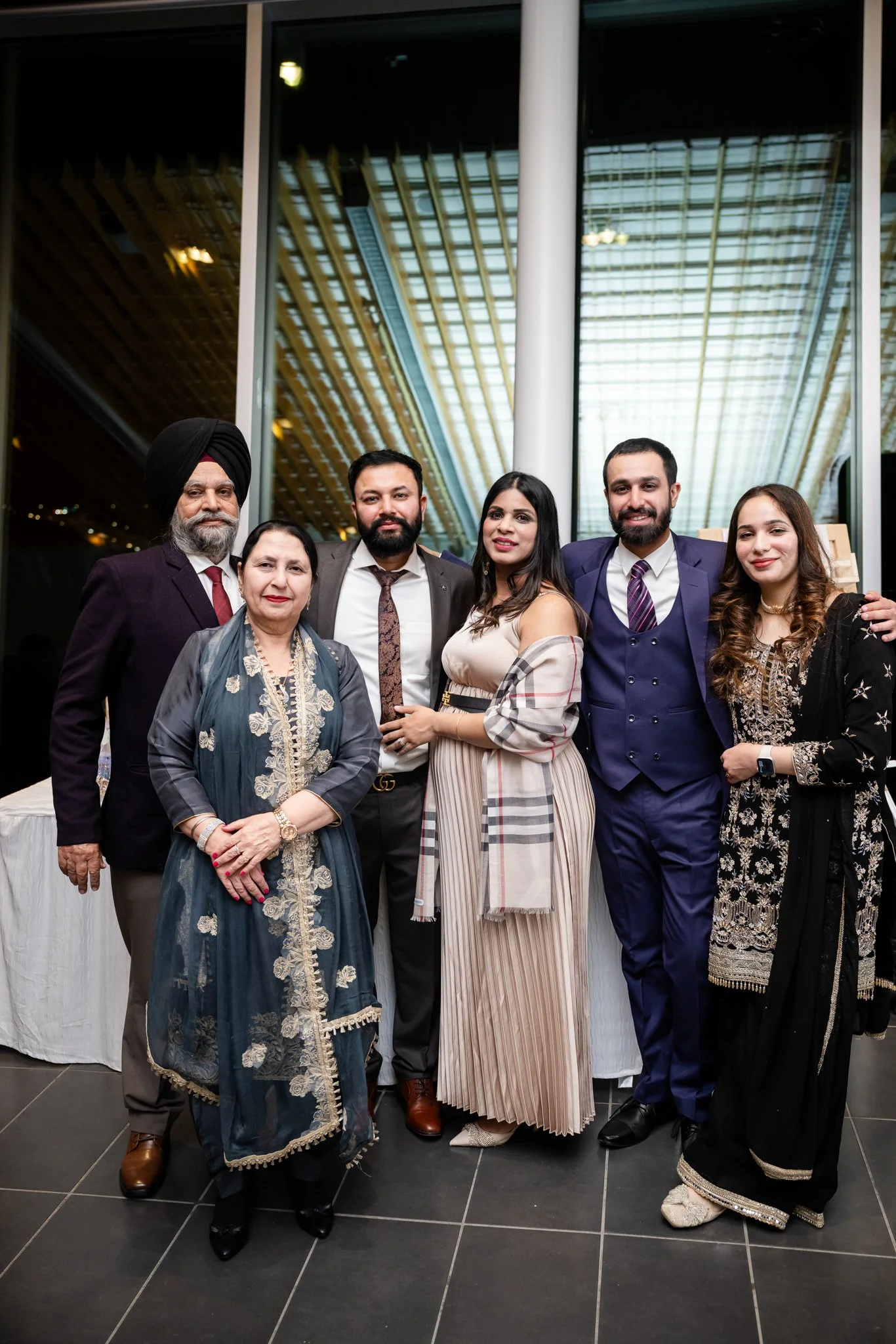 Group of seven people dressed in formal attire standing together indoors at an event, with a large window behind them showing a modern architectural ceiling.