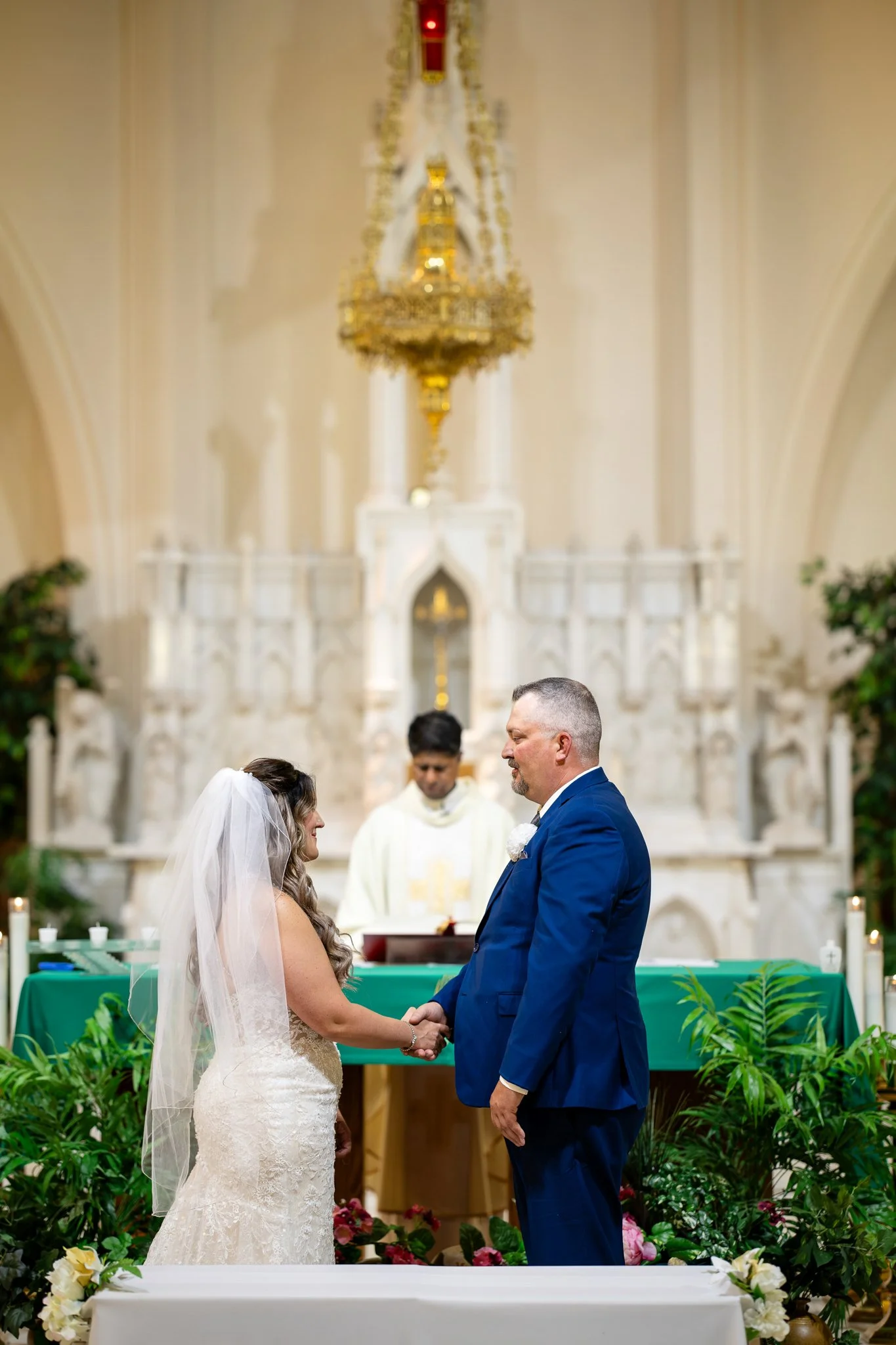 Bride and groom are holding hands during their wedding ceremony inside a church with an officiant and ornate altar in the background, surrounded by green plants and flowers.