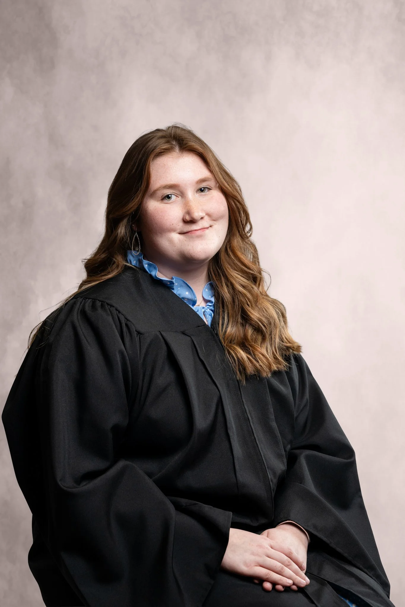 Young woman with long wavy auburn hair wearing a black graduation gown and a blue collared shirt, seated with hands resting on her lap, smiling softly, against a neutral background.