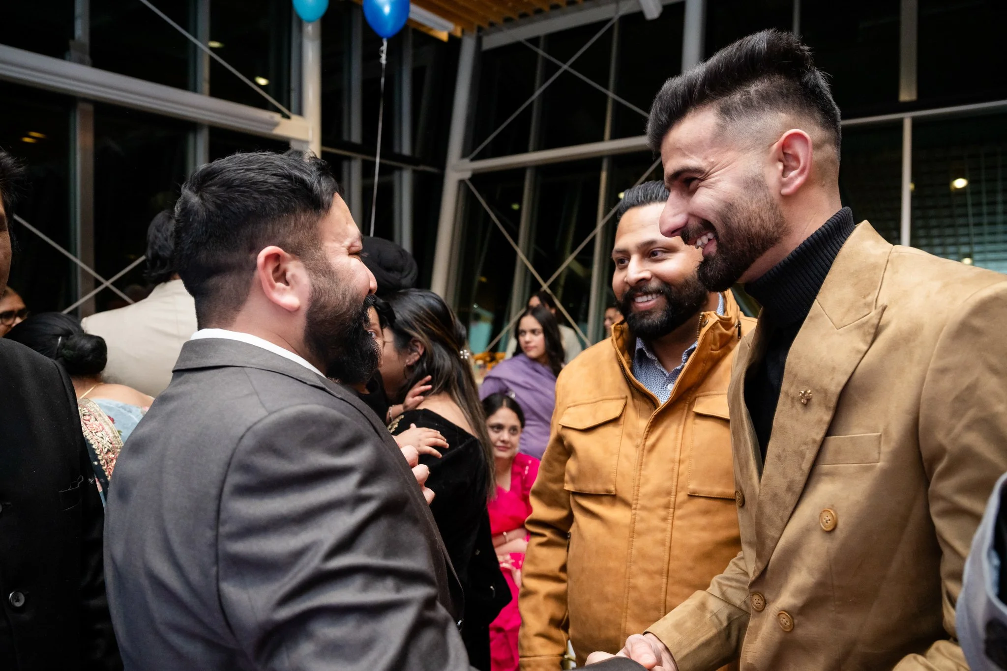 Three men smiling and shaking hands at a social event, with a woman in a pink dress and other guests in the background.
