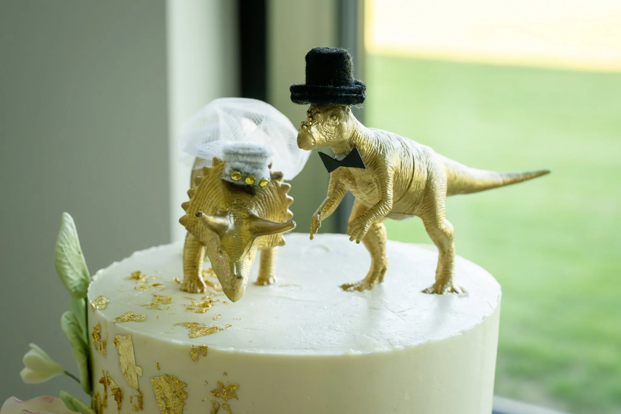 Dinosaur-themed wedding cake with two yellow dinosaur figures dressed as a bride and groom, wearing a white veil and black top hat with a bow tie, adorned with gold leaf details.