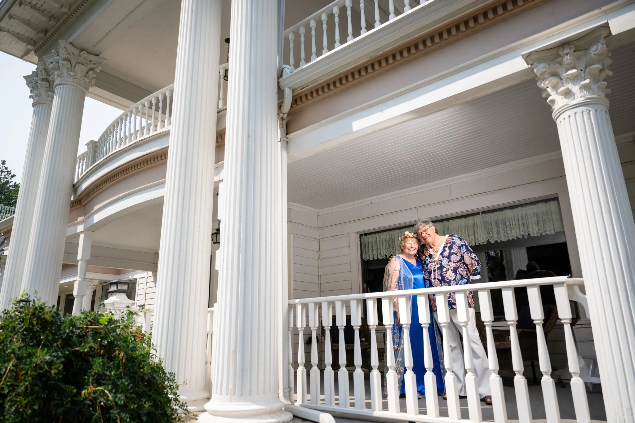 A smiling couple standing on a white porch of a large, elegant house with tall columns. The woman is wearing a blue dress with a floral sash and a flower crown. The man is wearing a patterned shirt and white pants.