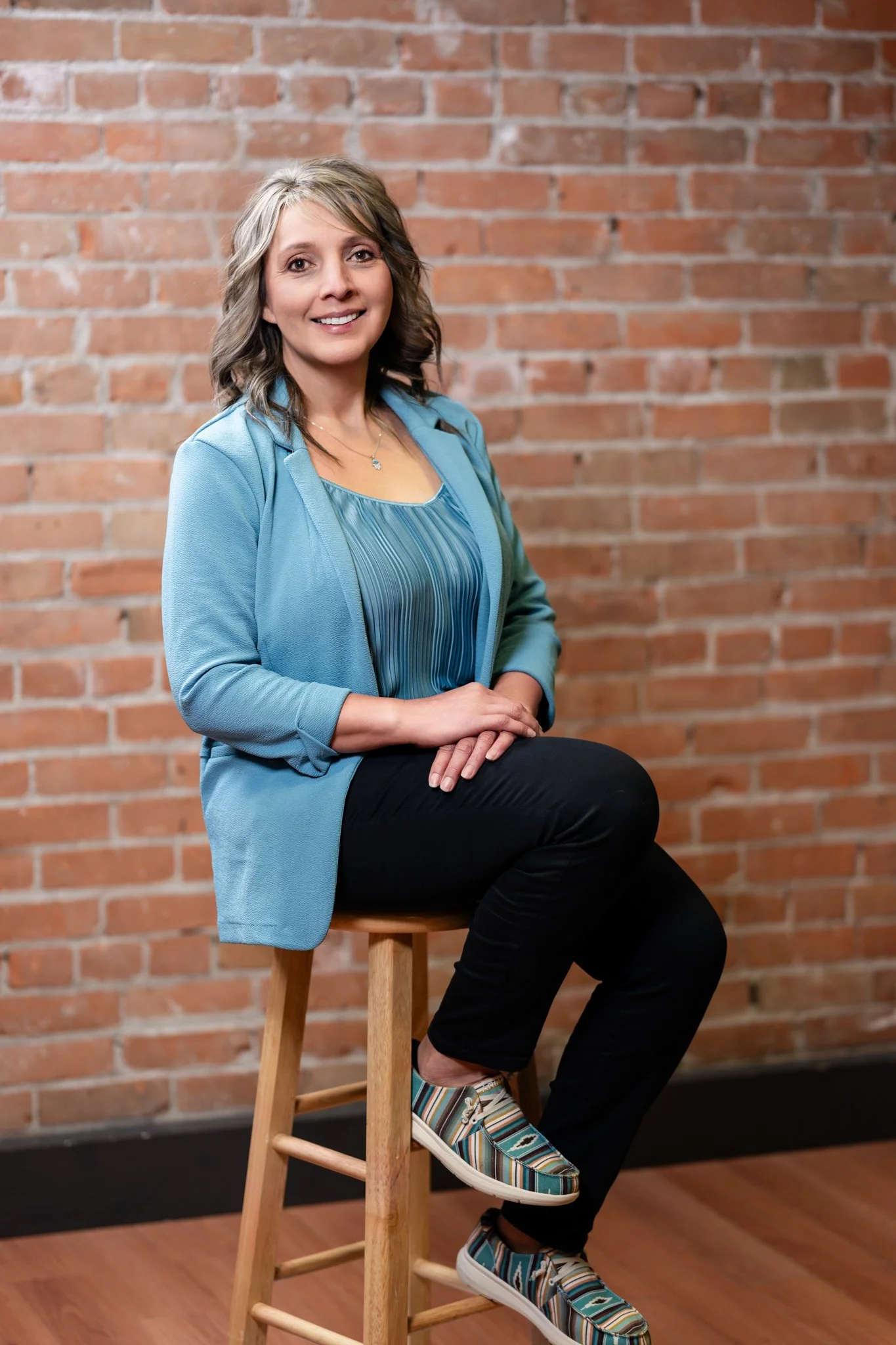 A woman with shoulder-length gray hair, smiling, sitting on a wooden stool against a brick wall. She is dressed in a blue blazer, blue patterned blouse, black pants, and colorful striped sneakers.