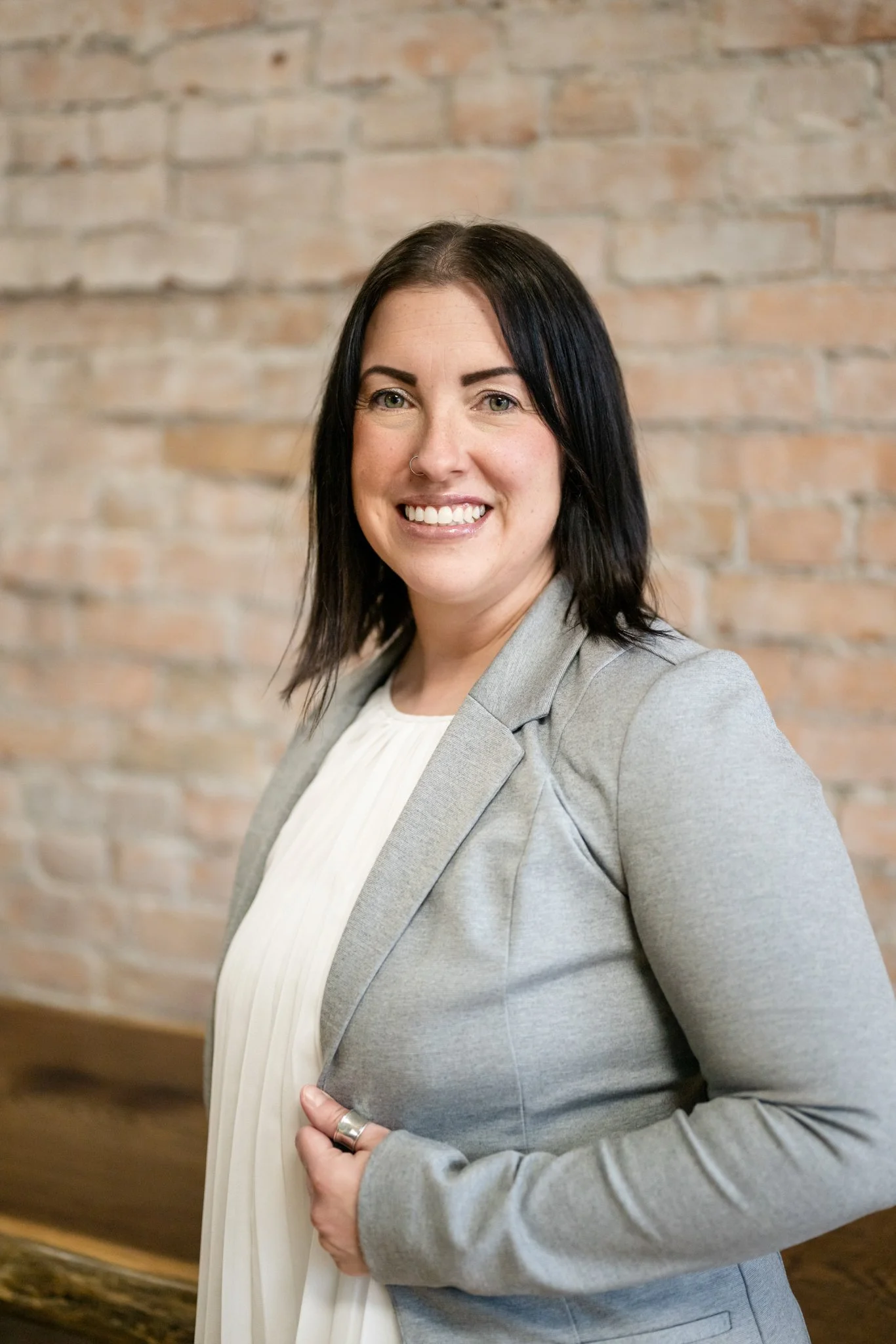 A woman with black hair, wearing a gray blazer and white blouse, smiling at the camera against a brick wall background.
