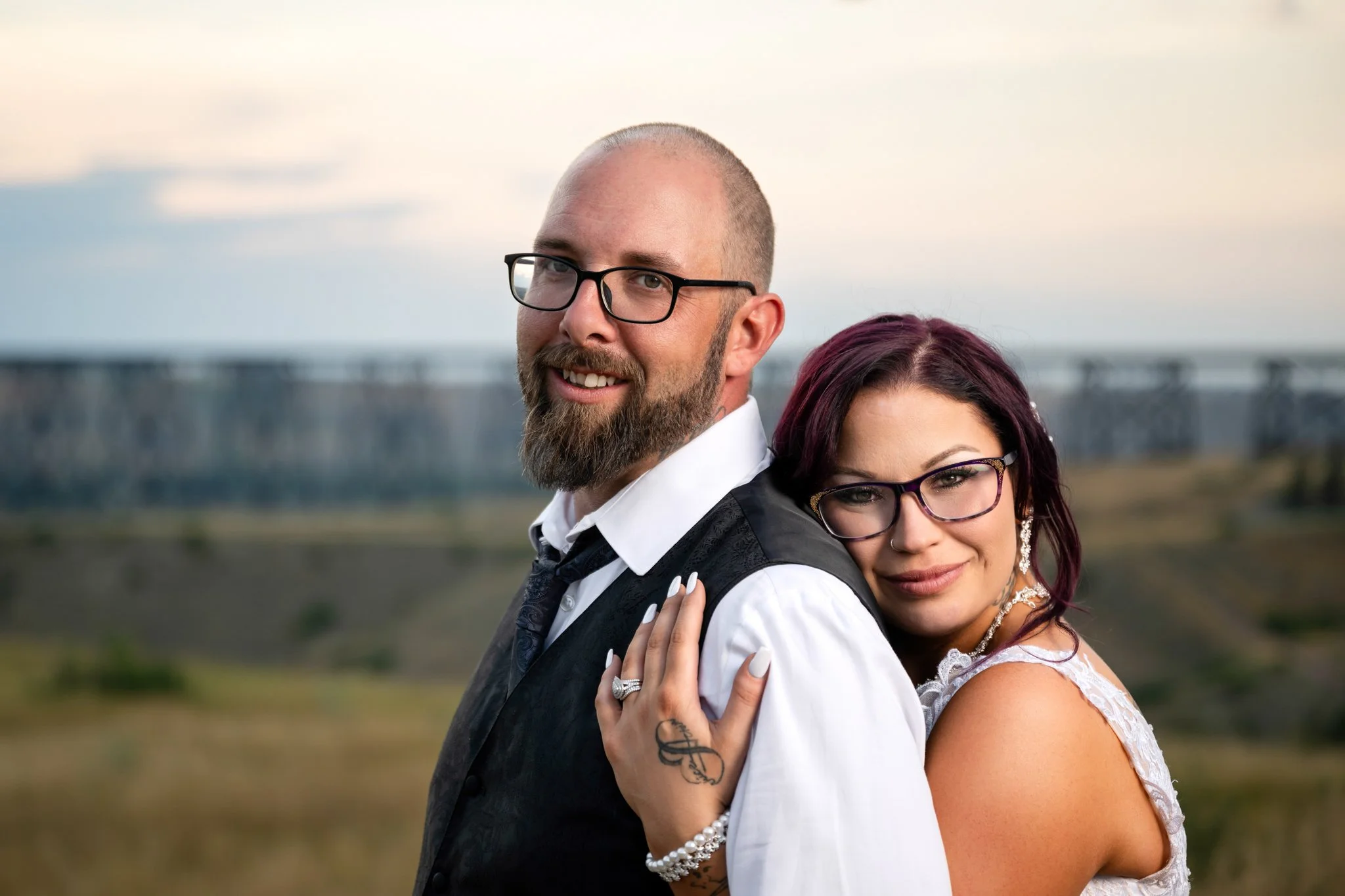 A man and woman are standing outdoors, close together, smiling. The man has a beard, glasses, and a shaved head, and is wearing a white shirt with a black vest. The woman has dark purple hair, glasses, and is wearing a white lace dress and jewelry. T