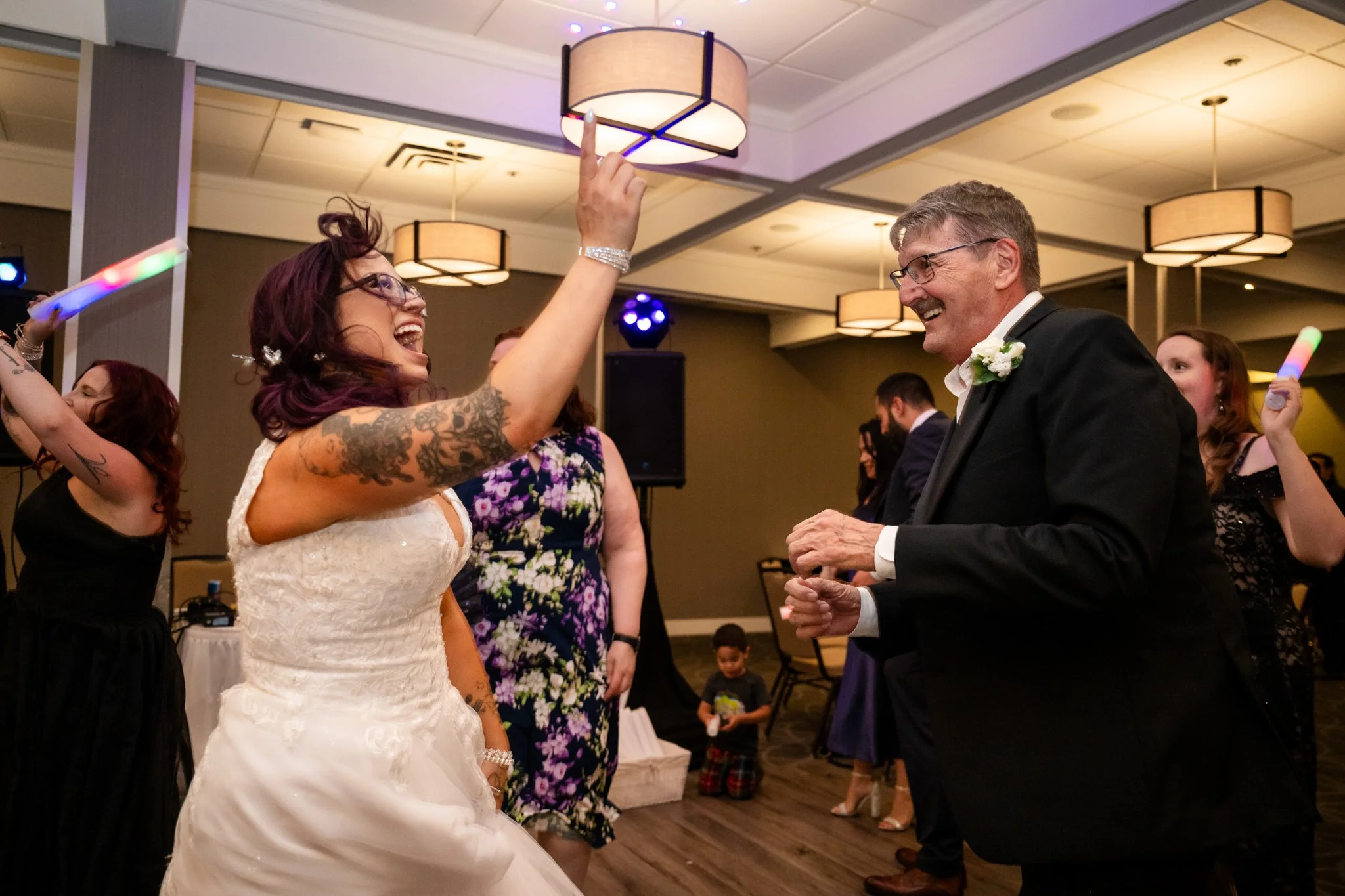 A bride and an older man are dancing and laughing at a wedding reception. The bride has tattoos on her arm, wears glasses, and is in a white wedding dress. The man is wearing a black tuxedo with a white shirt and a boutonniere. Other guests can be se