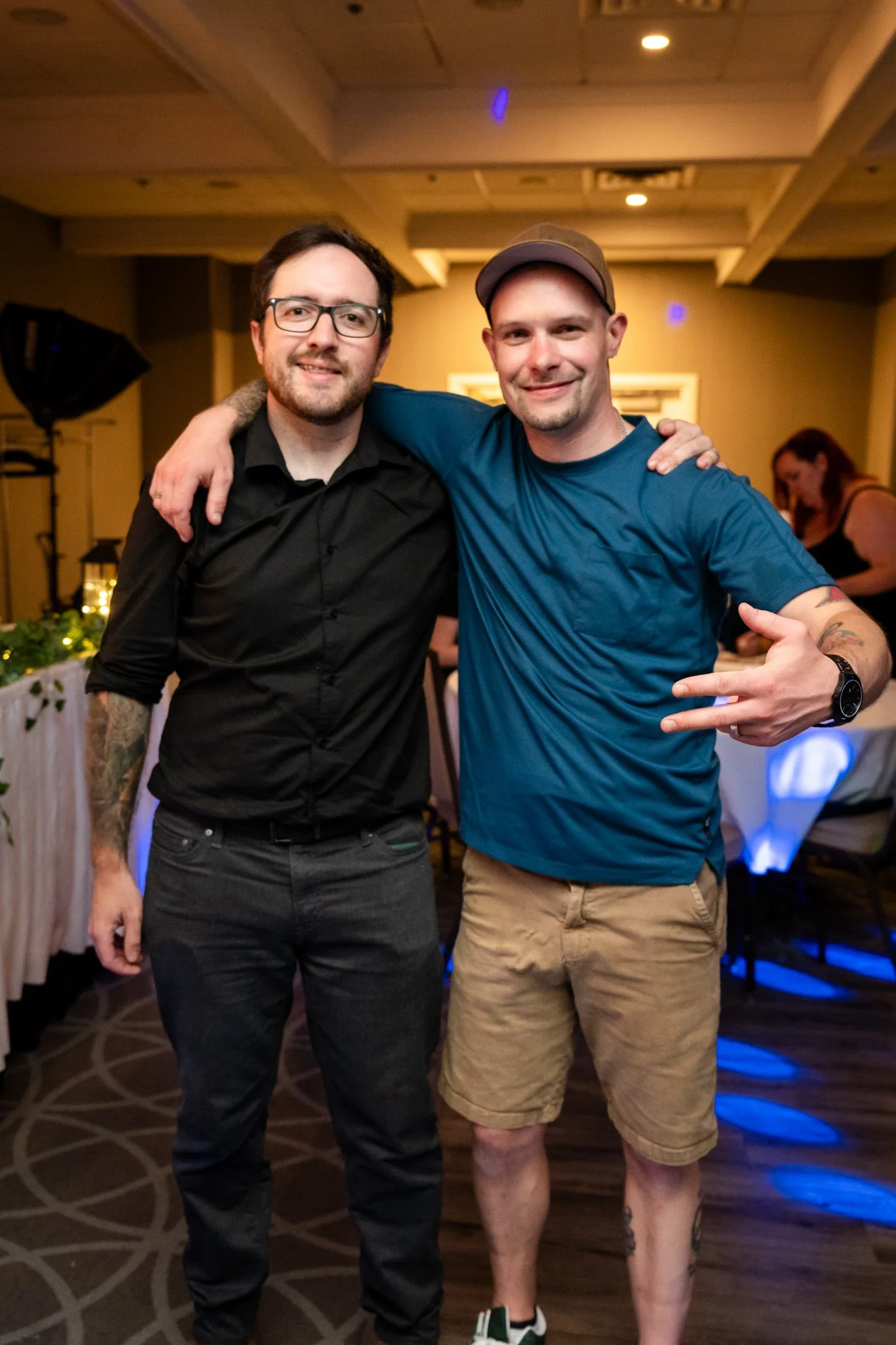 Two men standing closely together with arms around each other, smiling at a social gathering in an indoor venue. One man has dark hair, glasses, a black shirt, and tattoos on his arms. The other man wears a blue T-shirt, beige shorts, a cap, and has