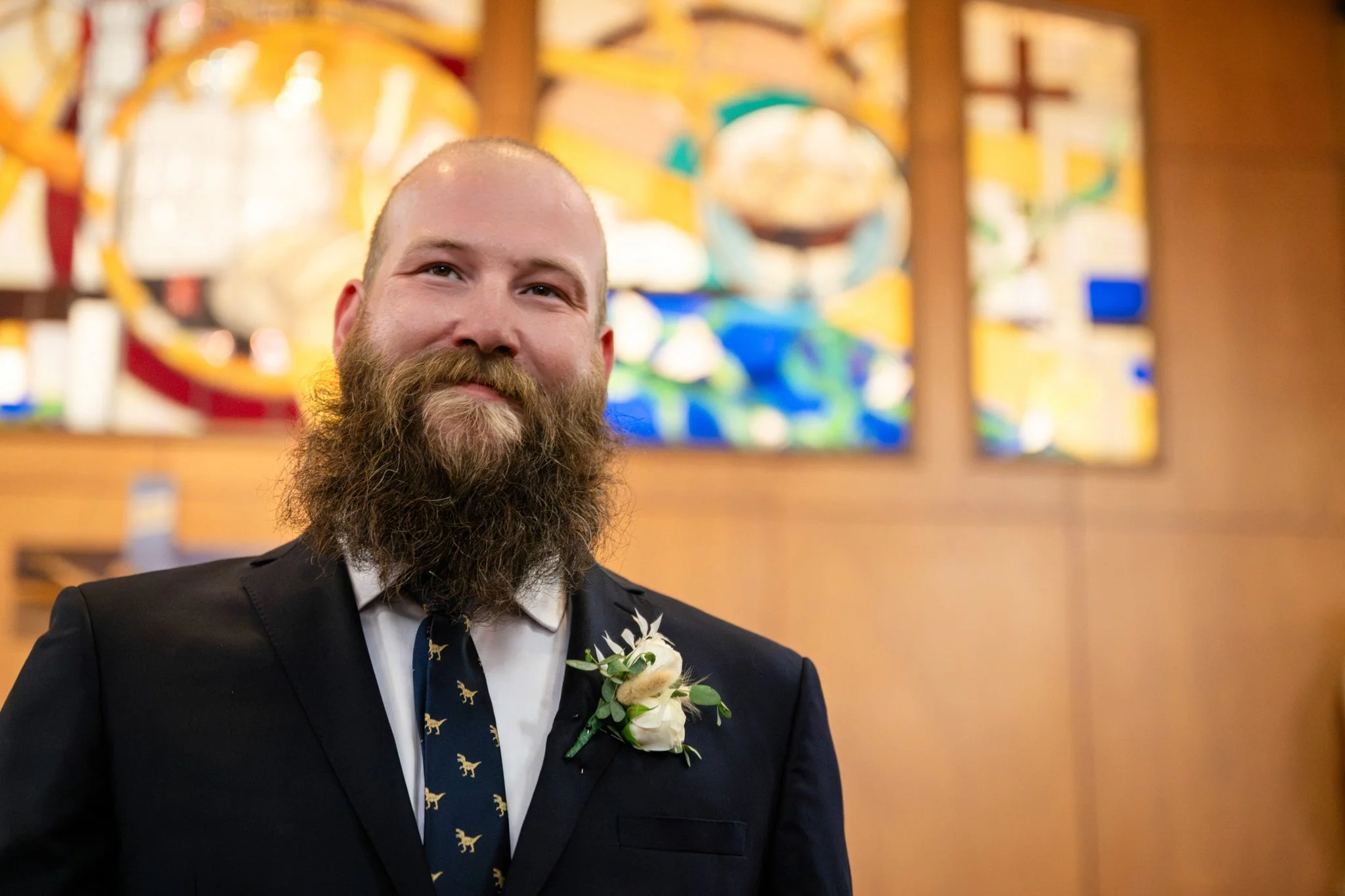 A man with a beard in a suit standing inside a church or chapel, with stained glass windows in the background.