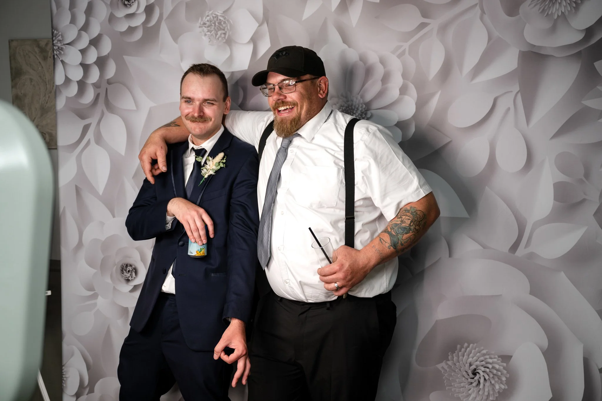 Two men smiling and posing with arms around each other, standing in front of a floral-themed wallpaper. One man is wearing a navy suit with a boutonniere, the other is dressed more casually in a white shirt, suspenders, and a hat with glasses, tattoo