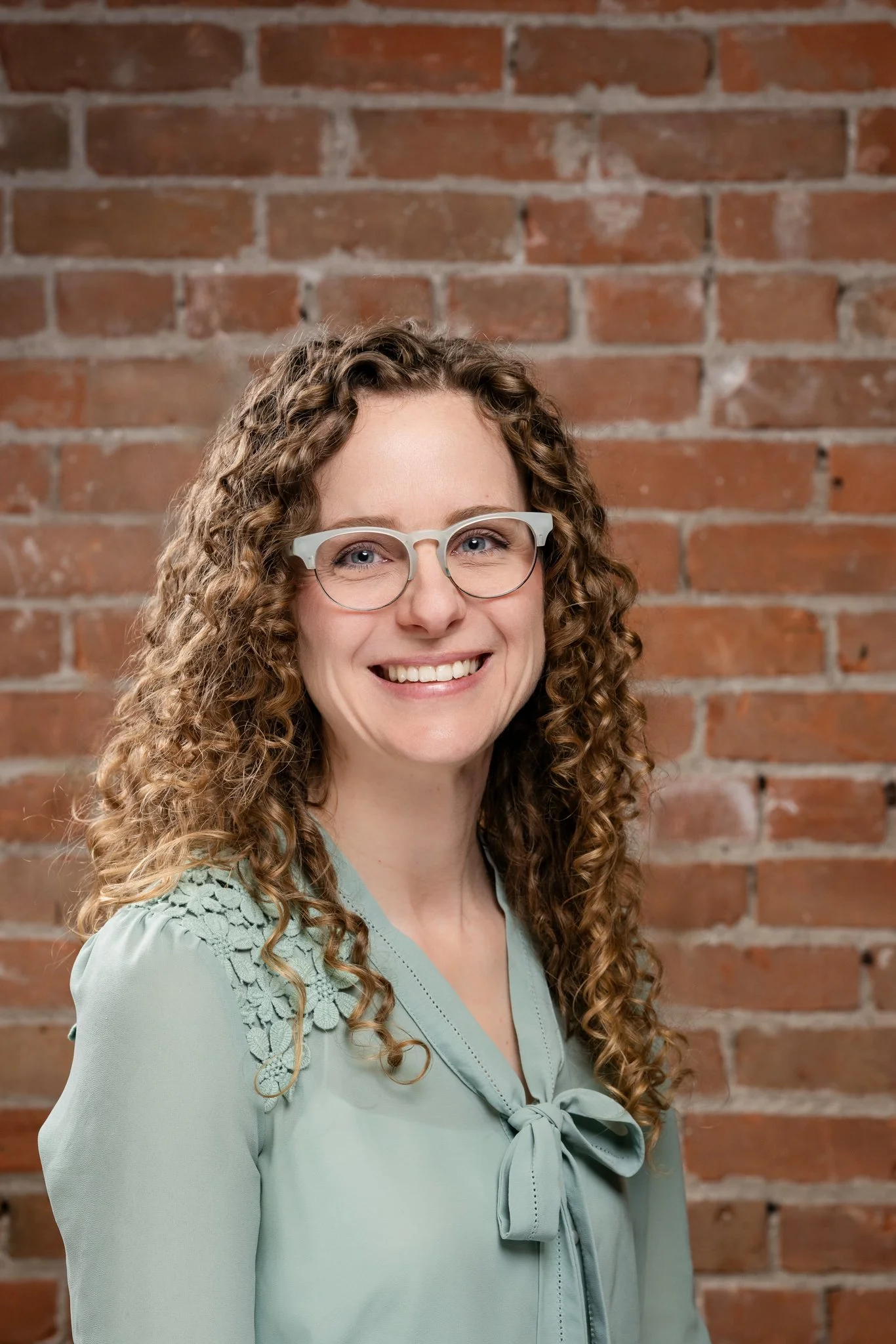 A woman with curly hair and glasses smiling, standing in front of a brick wall.