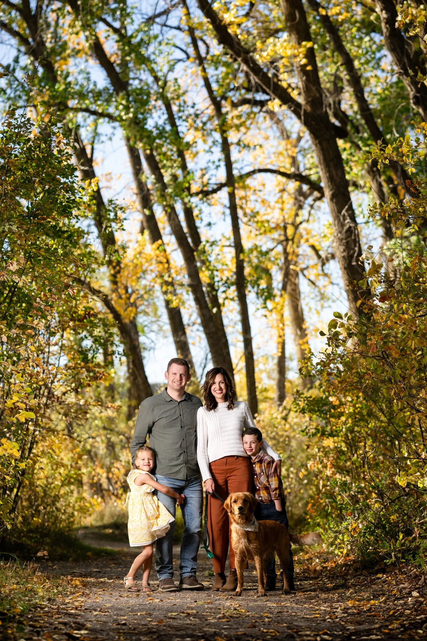 A family of four with a dog standing on a forest path surrounded by yellow and green autumn trees.