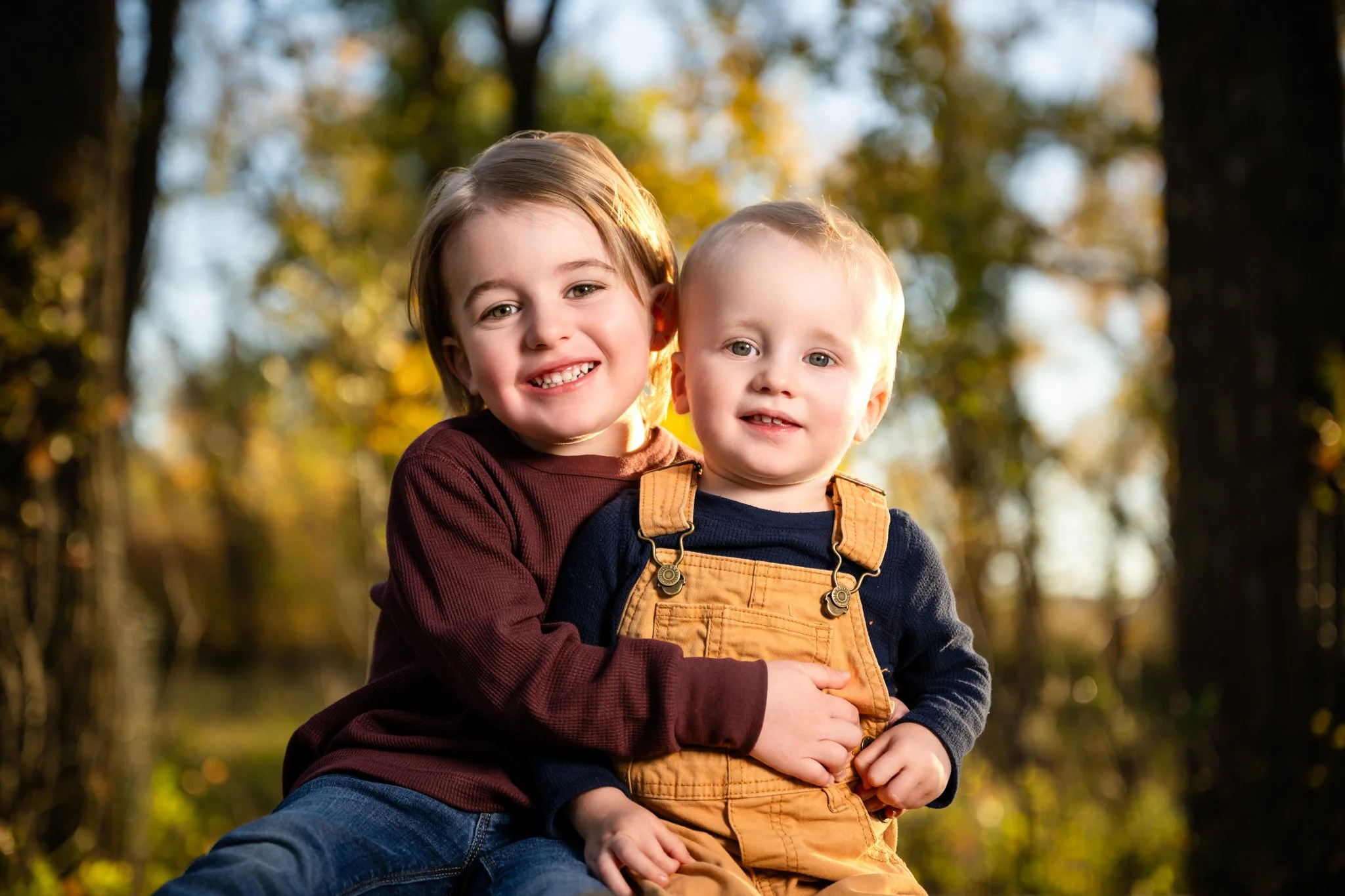 Two young children, a girl and a boy, sitting outdoors during autumn. They are smiling and hugging each other, with fall foliage in the background.