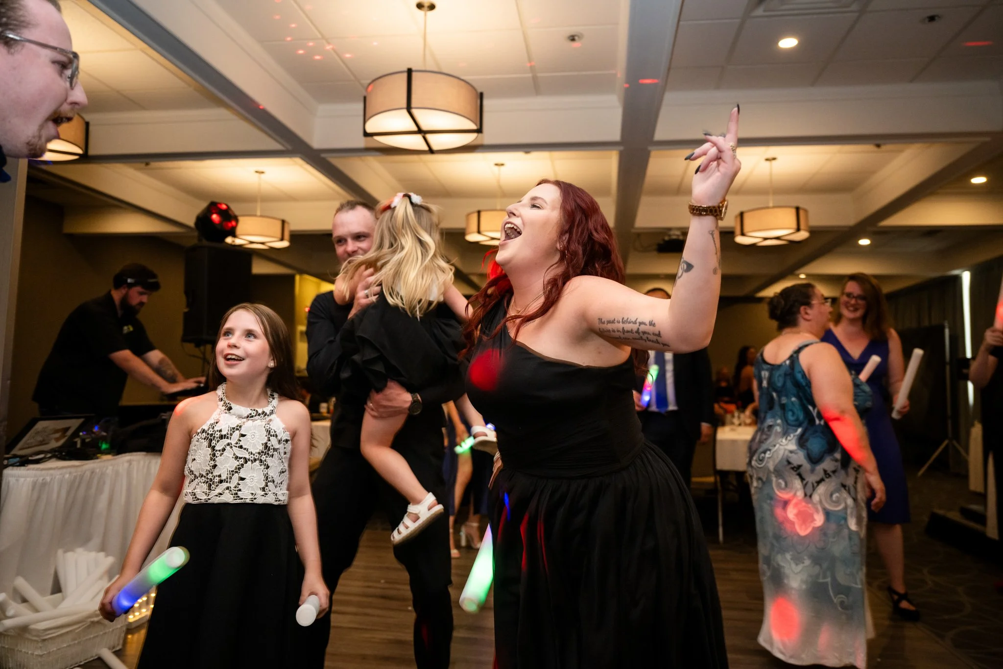People dancing and celebrating at an indoor party, with a woman singing and holding up her hand, children holding glow sticks, and others smiling and enjoying the lively atmosphere.