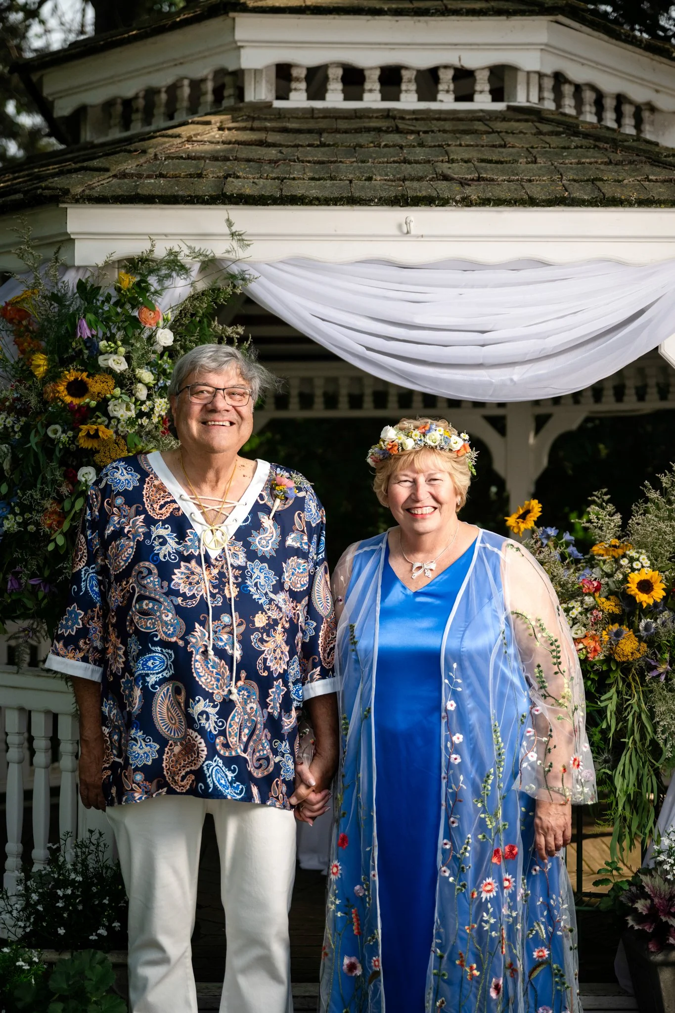An elderly couple standing hand-in-hand under a decorated gazebo, smiling at the camera, surrounded by colorful flowers, at a garden wedding or celebration.