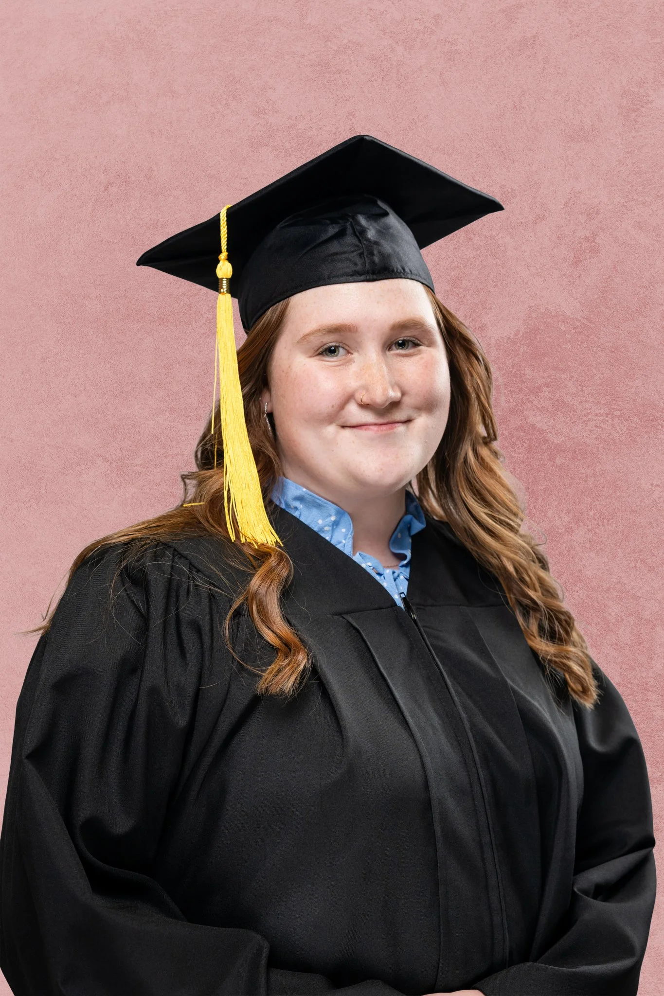 Young woman in a graduation cap and gown smiling against a pink background.