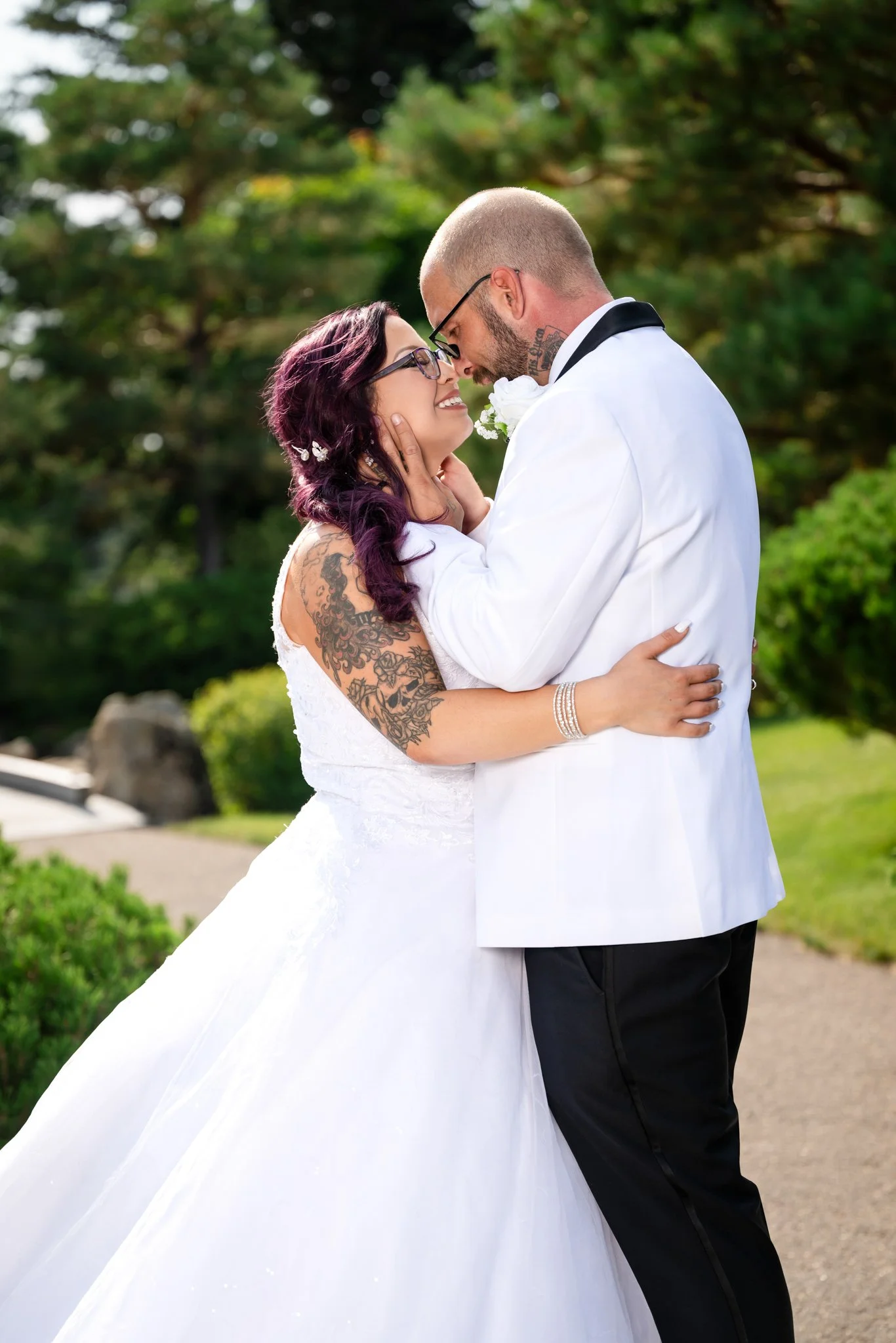 A bride and groom embrace outdoors during their wedding, smiling and touching foreheads, with green trees and bushes in the background.