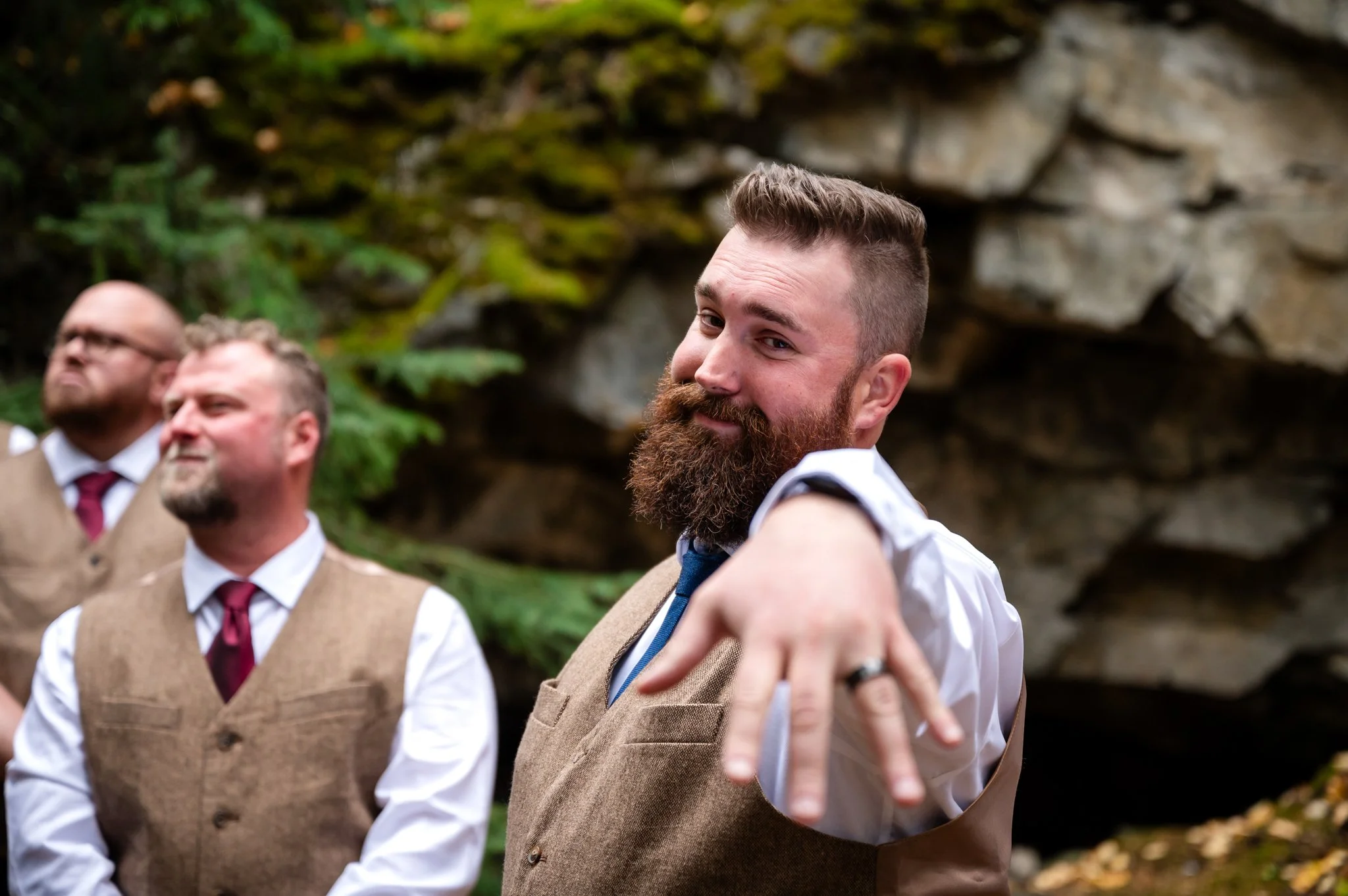 A man with a beard and mustache pointing at the camera, wearing a white shirt, tie, and brown vest, with two other men in similar attire in the background outdoors near rocky scenery.