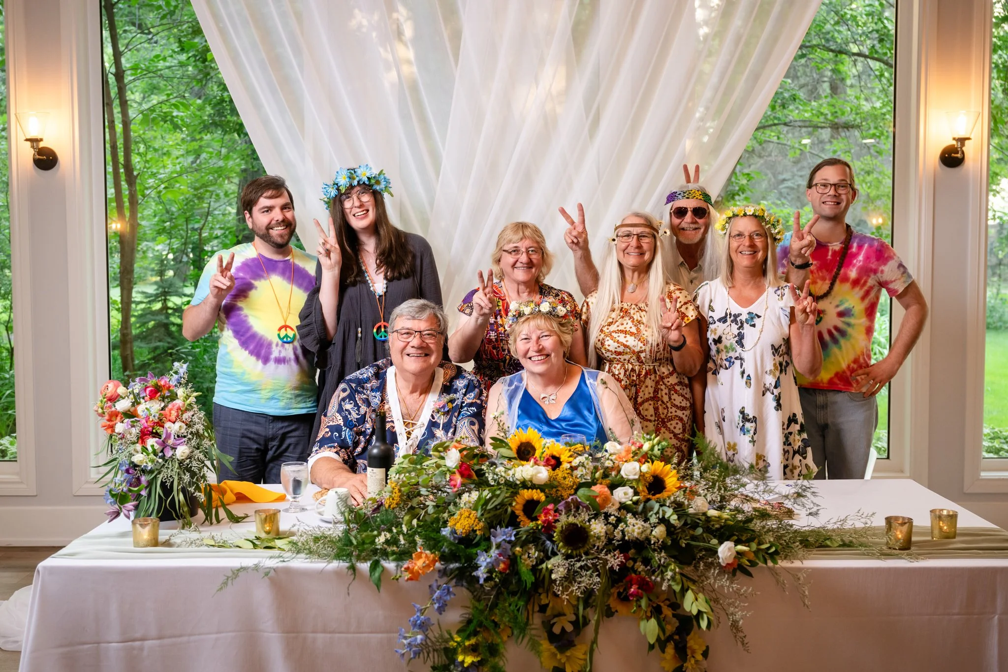 A group of people at a celebration dressed in colorful, tie-dye clothing and accessories with peace sign necklaces. They are making peace signs and smiling, with a decorated floral table, large window, and green outdoor scenery in the background.