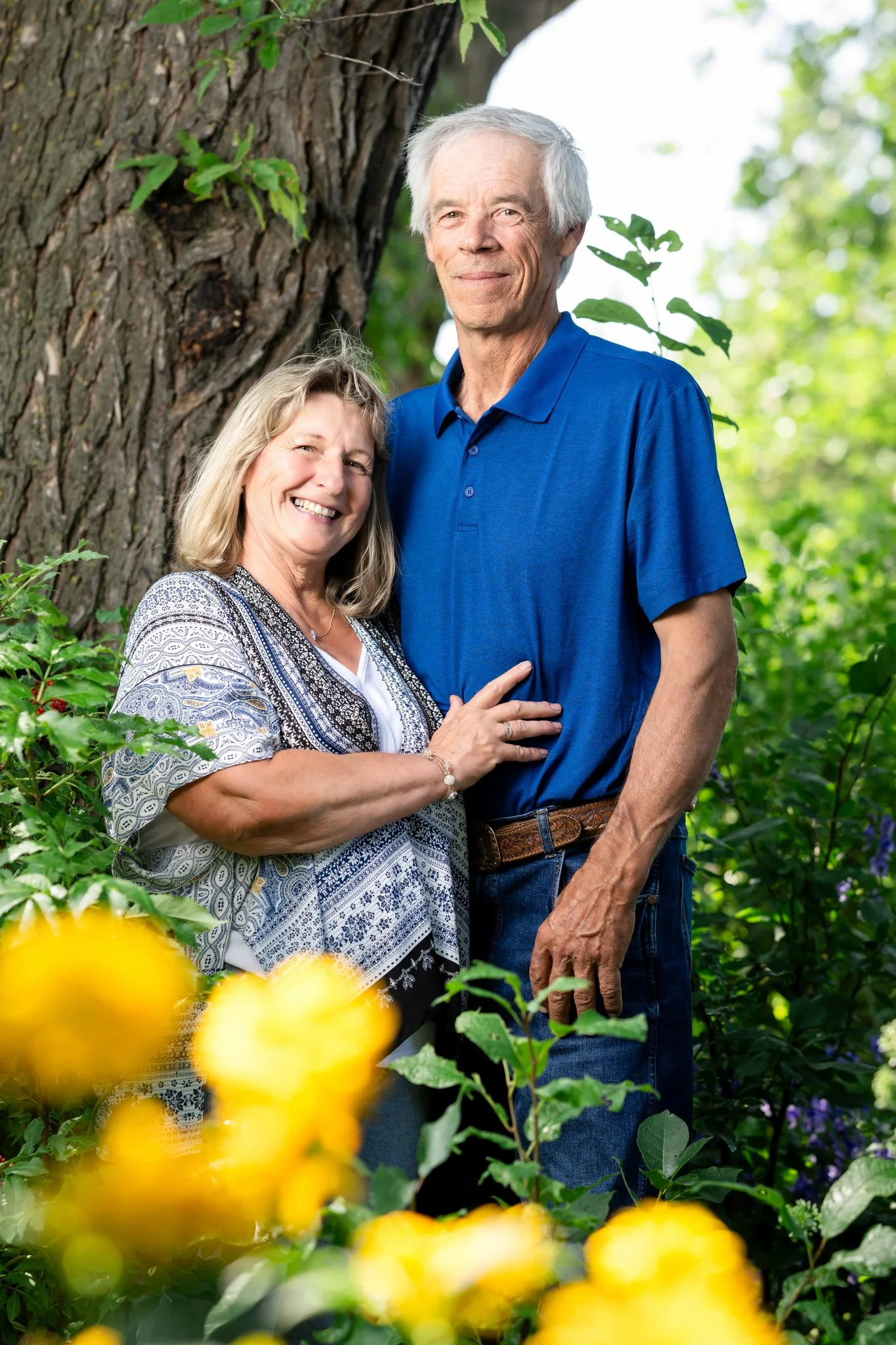 A smiling woman and an older man standing close together outdoors in front of a tree, surrounded by green foliage and yellow flowers.
