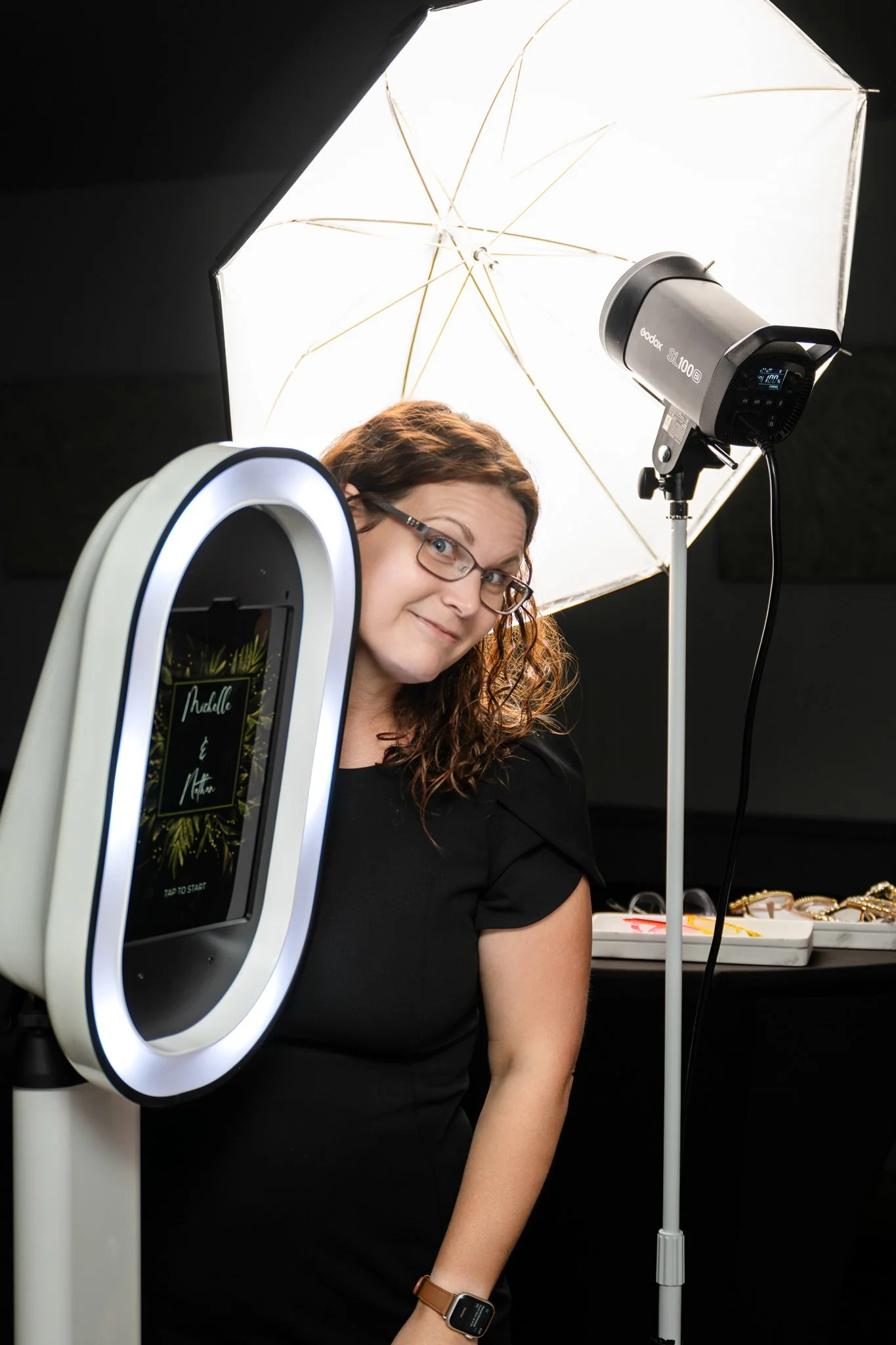 A woman with curly brown hair, glasses, and wearing a black shirt, stands in a photo booth with various photography equipment, including an umbrella light, a professional camera, and a mobile phone, displaying a welcome screen with names Madelle and