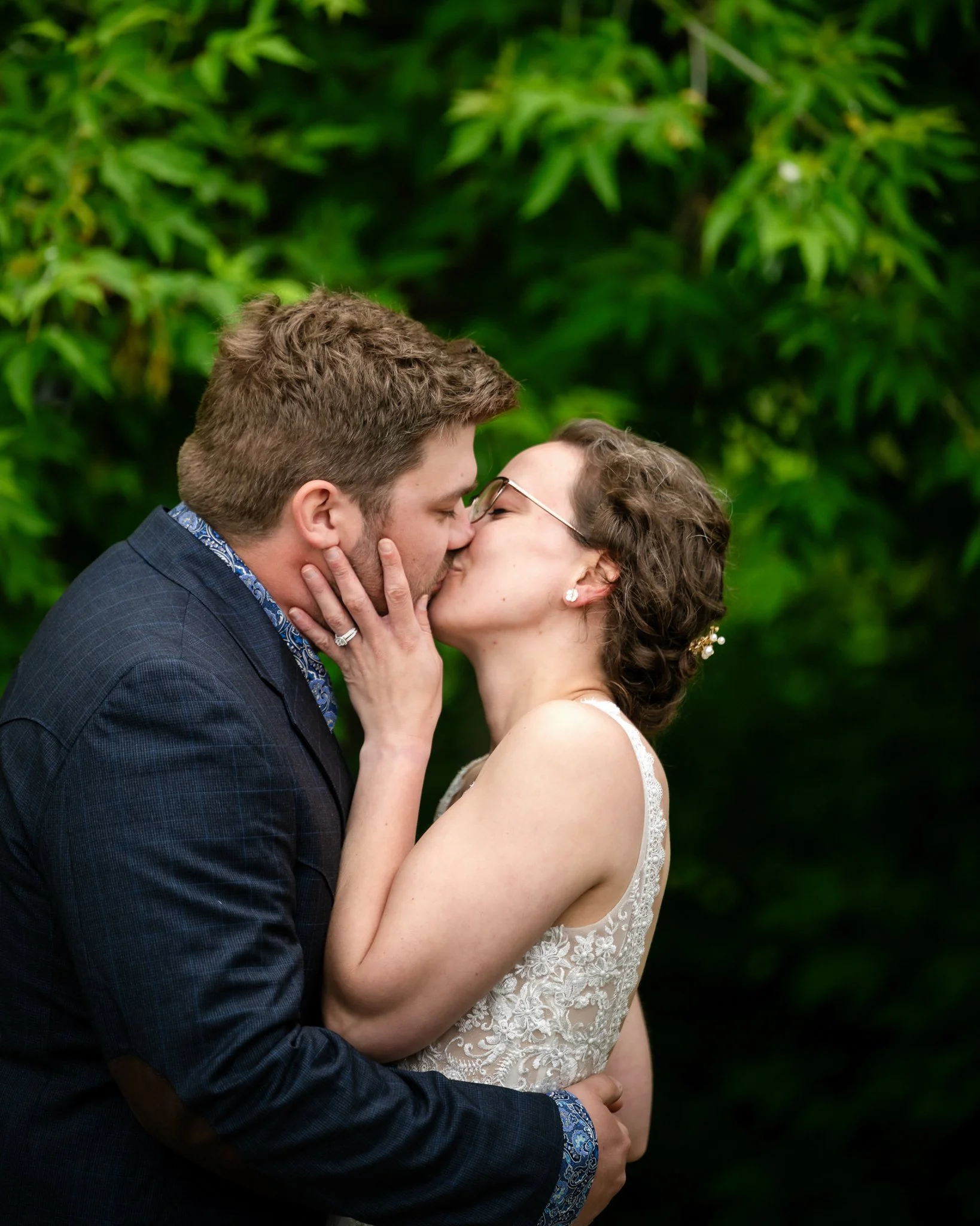 A newlywed couple sharing a kiss outdoors, the groom in a dark suit and the bride in a lace wedding dress, with green foliage in the background.