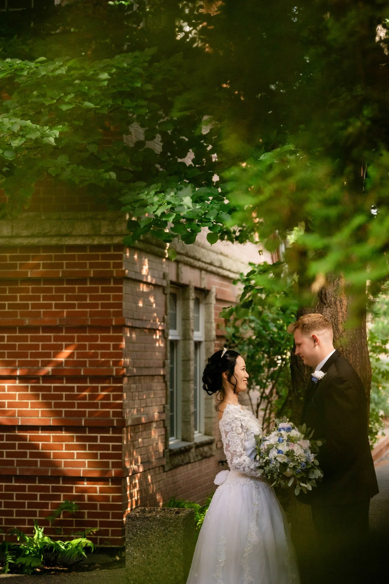 A bride and groom share a moment outdoors, standing close to each other beside a large tree, with greenery and a brick building in the background.