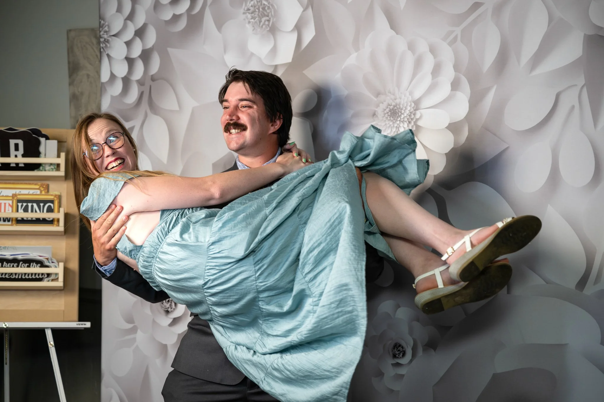 A man dressed in a dark suit and blue shirt holds a woman in a light blue dress. The woman has red hair, glasses, and is smiling as she is lifted off the ground. They are in a room with a large, white floral patterned wallpaper background. There is a