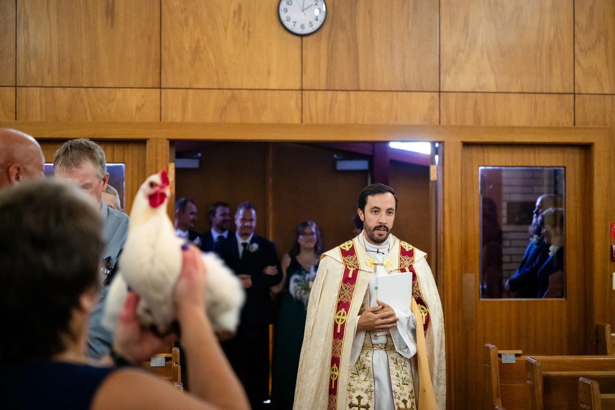 A priest standing in a church during a wedding ceremony, holding a book, with guests seated and a woman holding a chicken in the foreground.