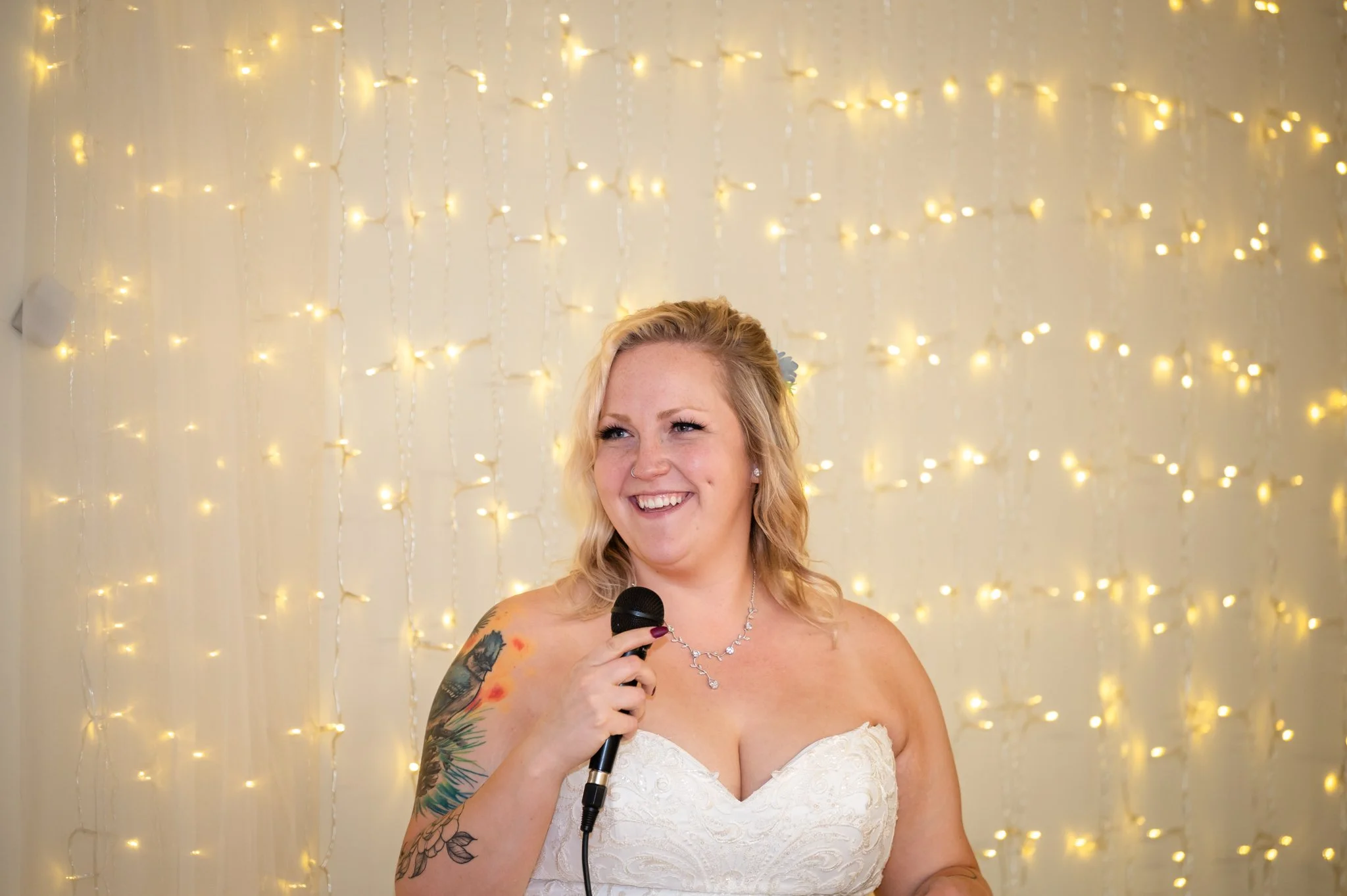 A smiling woman in a wedding dress holding a microphone, standing in front of a backdrop with string lights.