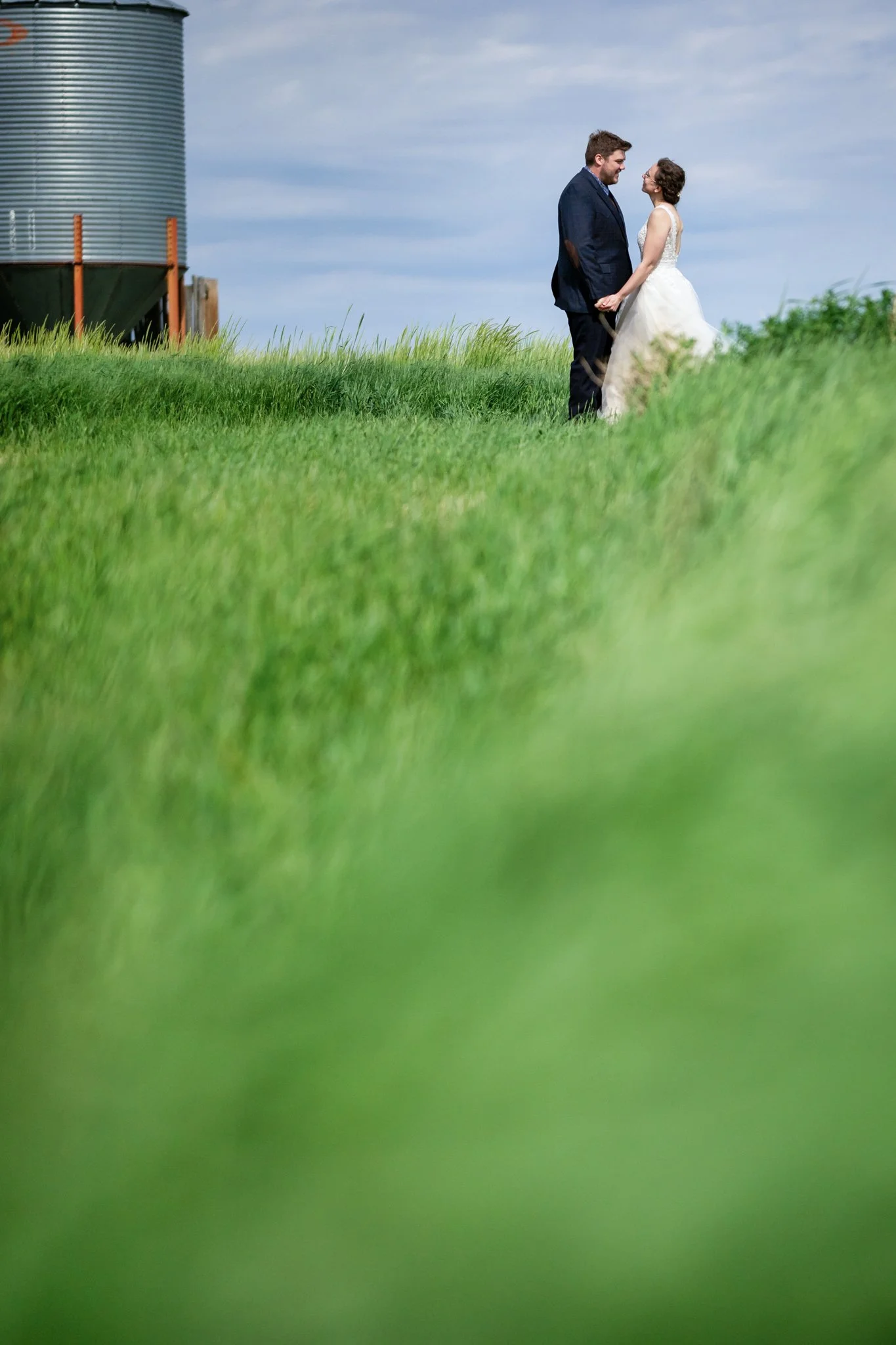 A couple dressed in wedding attire, holding hands and gazing at each other in a green grassy field with a large metal silo in the background.