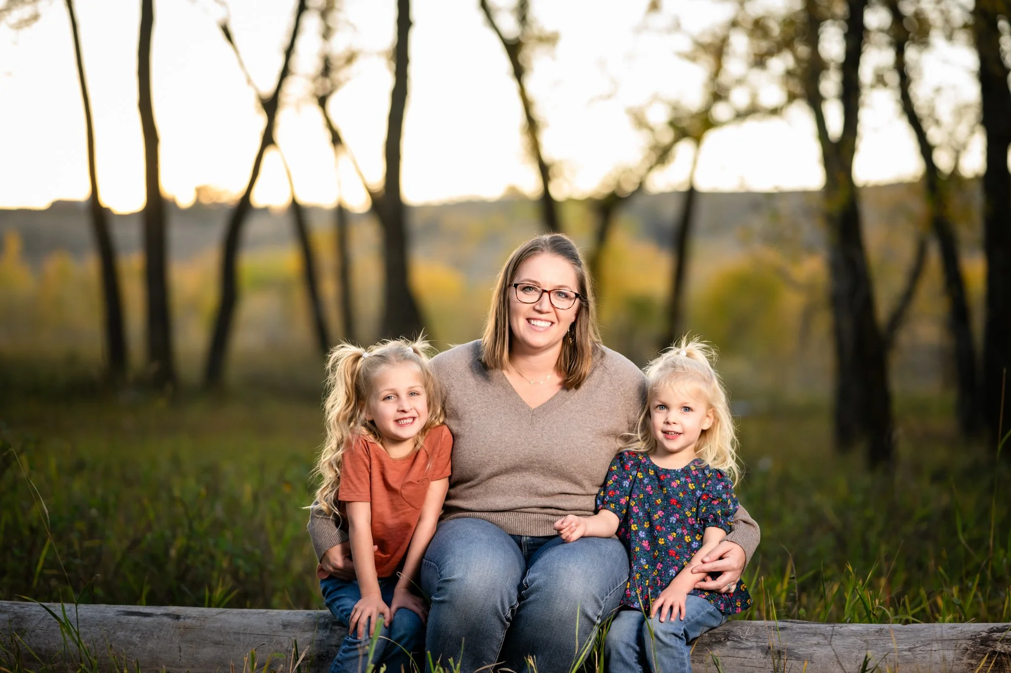 A woman sitting on a log outdoors with two young girls, smiling, during sunset in a wooded area.