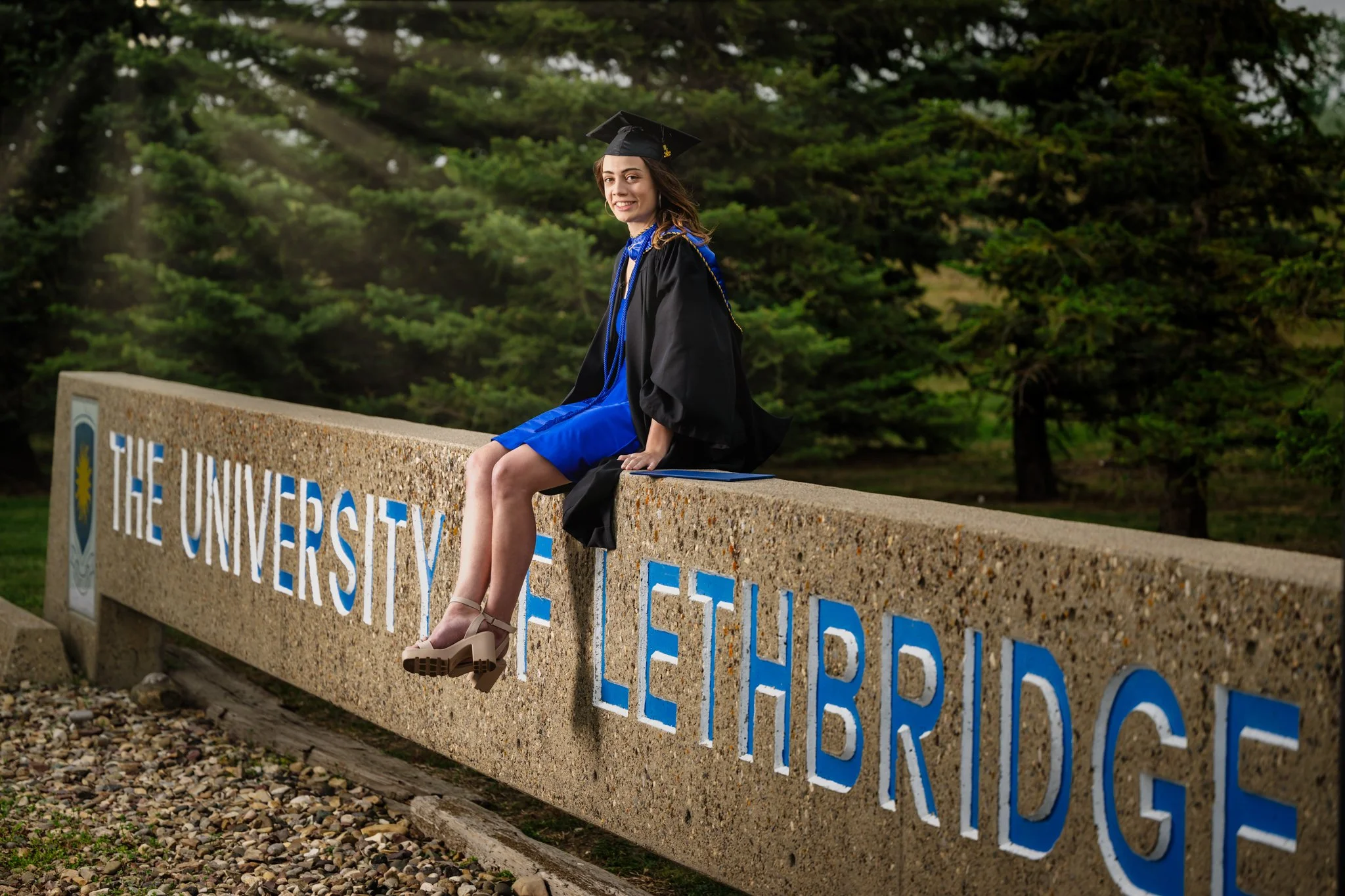 A woman in a graduation cap and gown sitting on a stone sign that reads "The University of Lethbridge," with trees and greenery in the background.