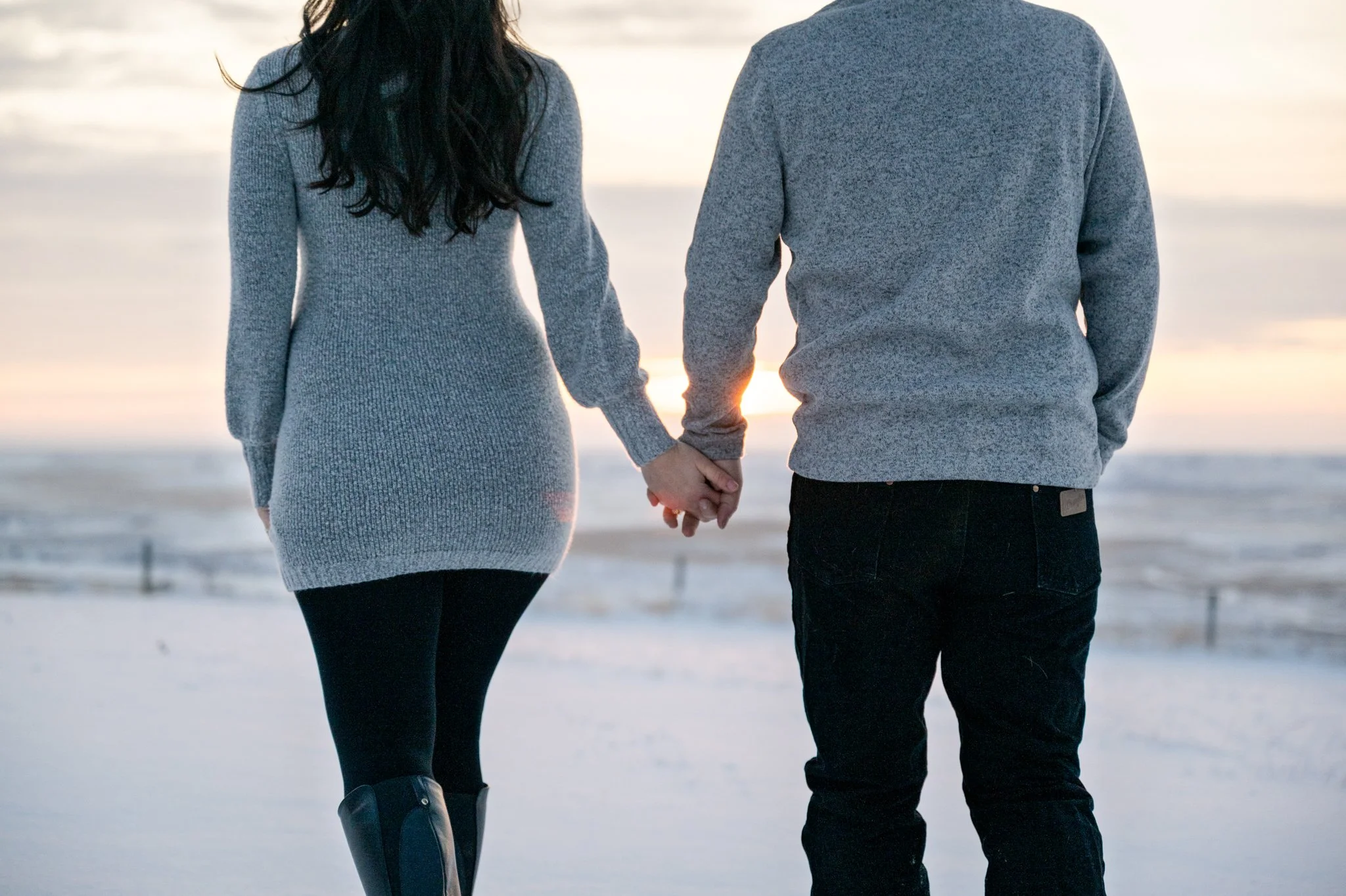 A couple holding hands, walking on a snowy beach at sunset, with the ocean in the background.