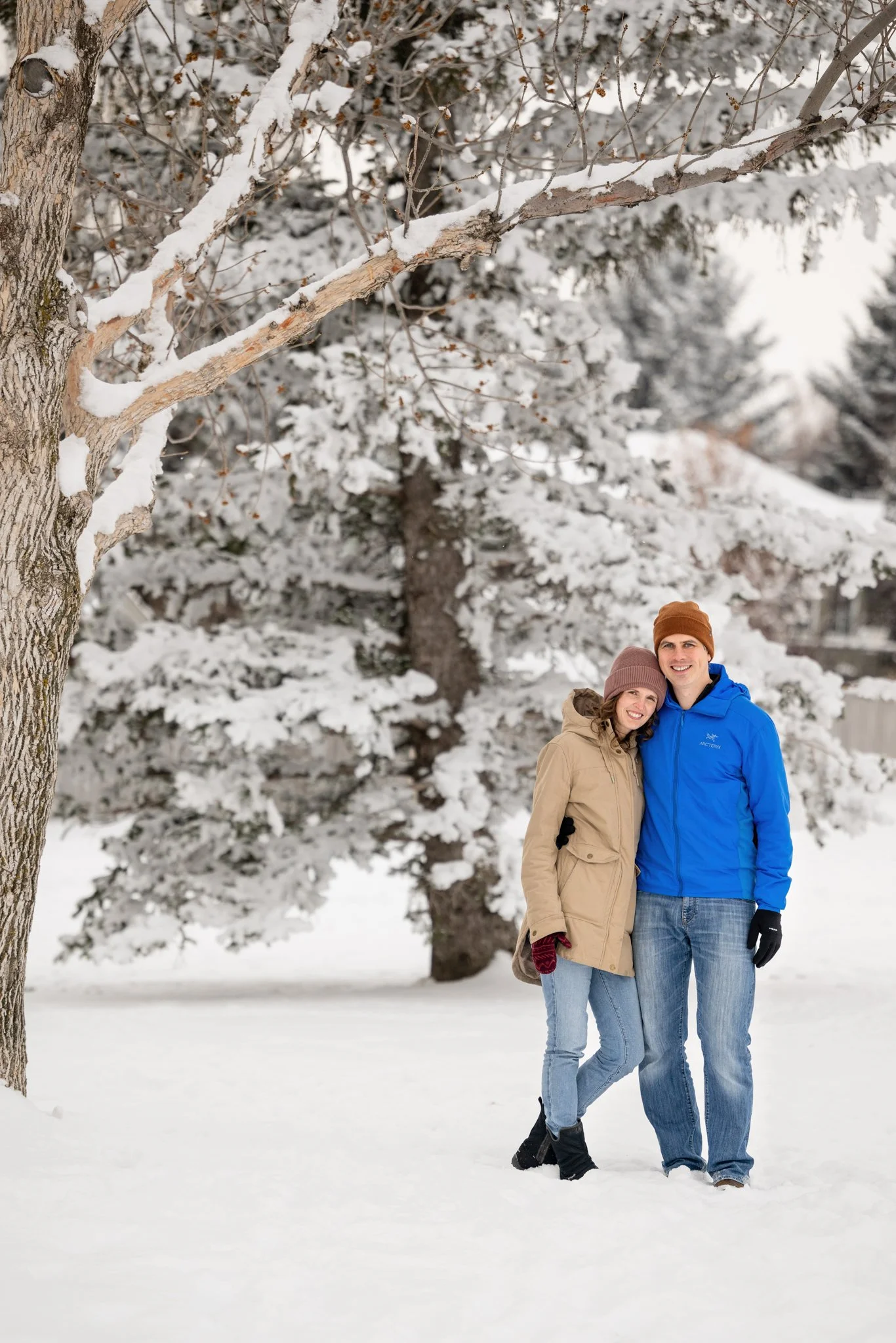 A couple standing on snow-covered ground, smiling, bundled in winter clothing, with snow-covered trees in the background.