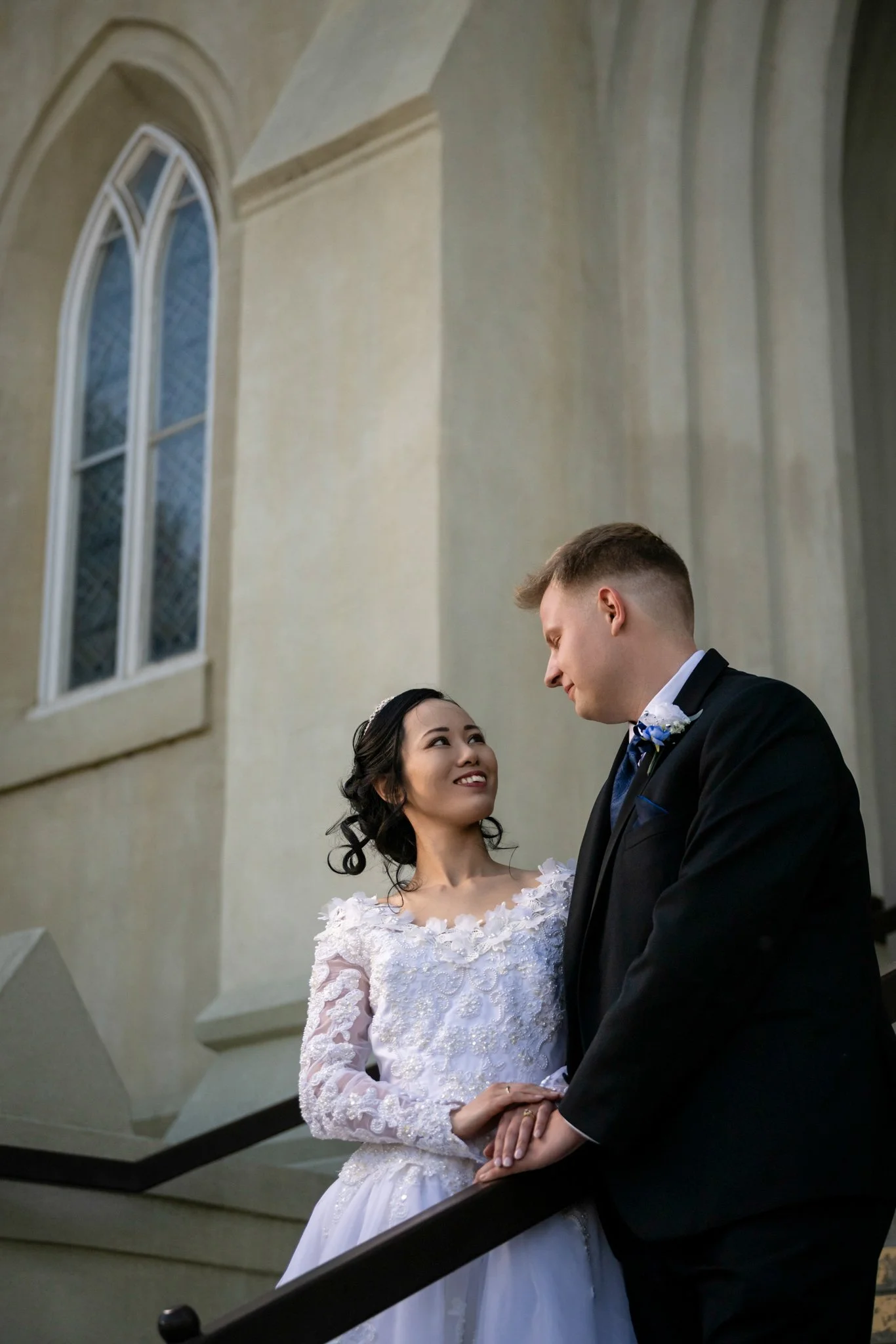A bride and groom standing on a staircase, gazing into each other's eyes inside a church or cathedral.