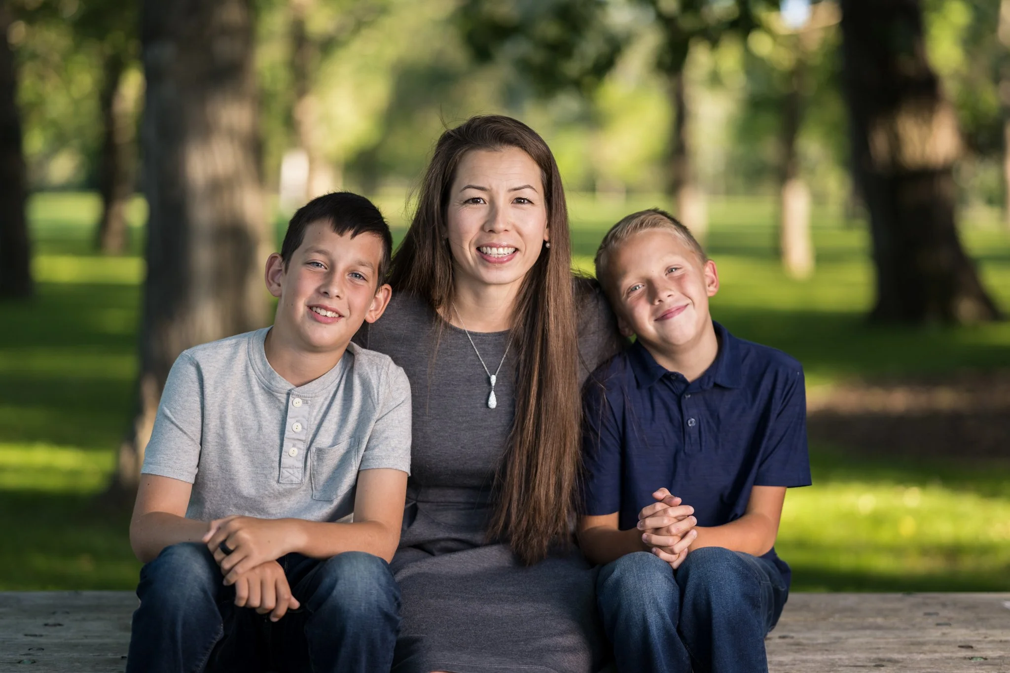 A woman sitting outdoors on a wooden bench with two boys, smiling at the camera, surrounded by trees and greenery.