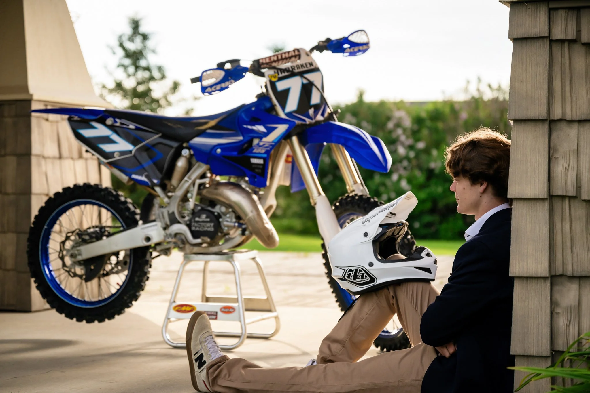 A young man sitting on the ground leaning against a wooden corner wall, with a blue and white motocross bike on a stand nearby and a white motorcycle helmet resting on his knee, outdoors during daytime.