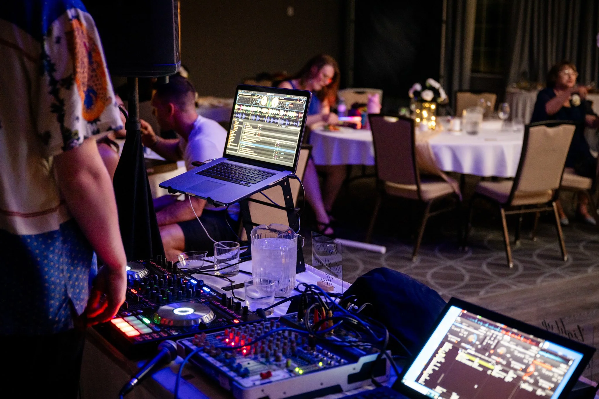 DJ setup with laptops, mixers, and DJ controllers at an event with people sitting at tables in the background.