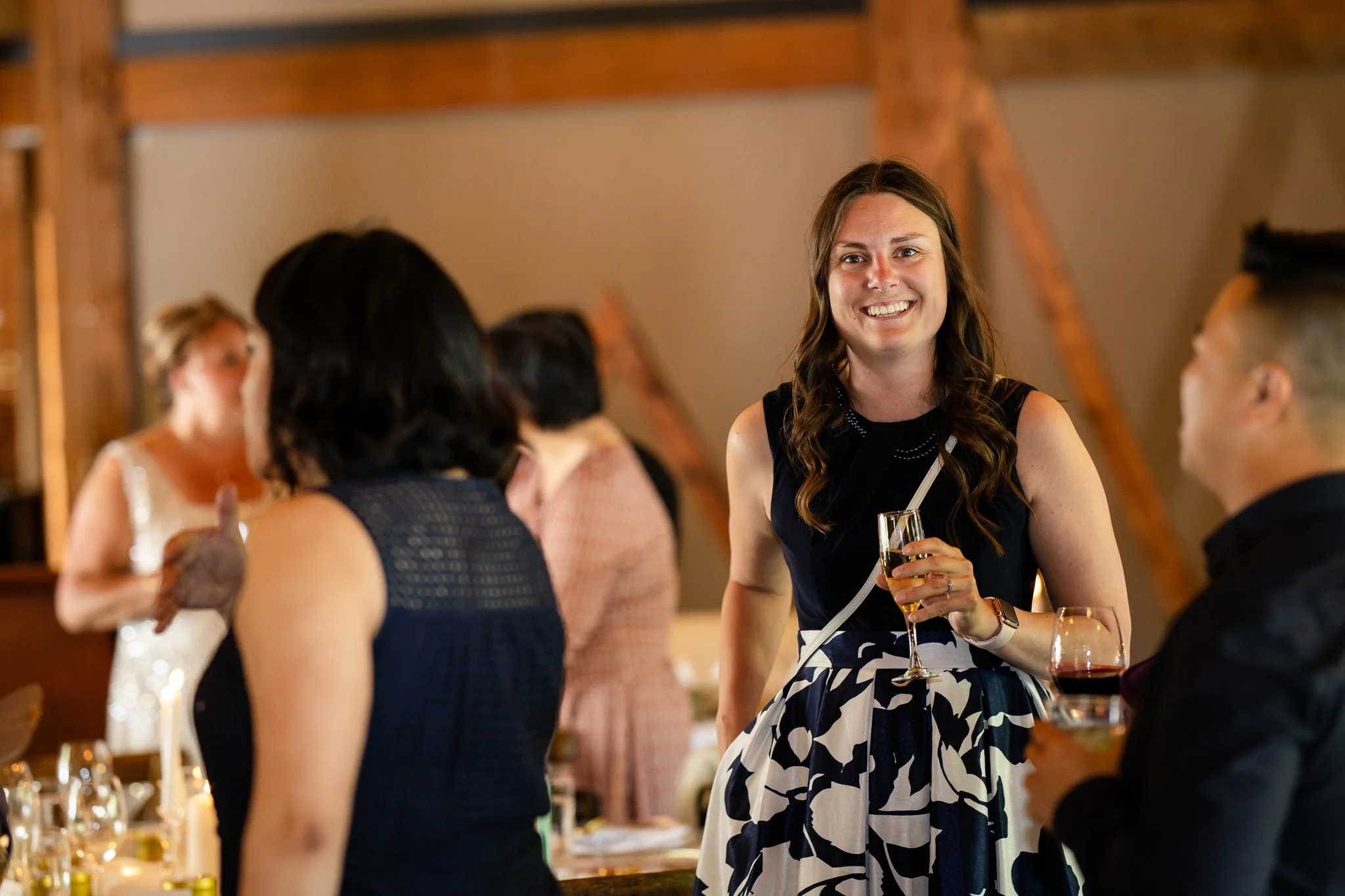 People at a social gathering, with a woman smiling and holding a glass of champagne, engaged in conversation with another guest, in a warmly lit room with wooden beams.